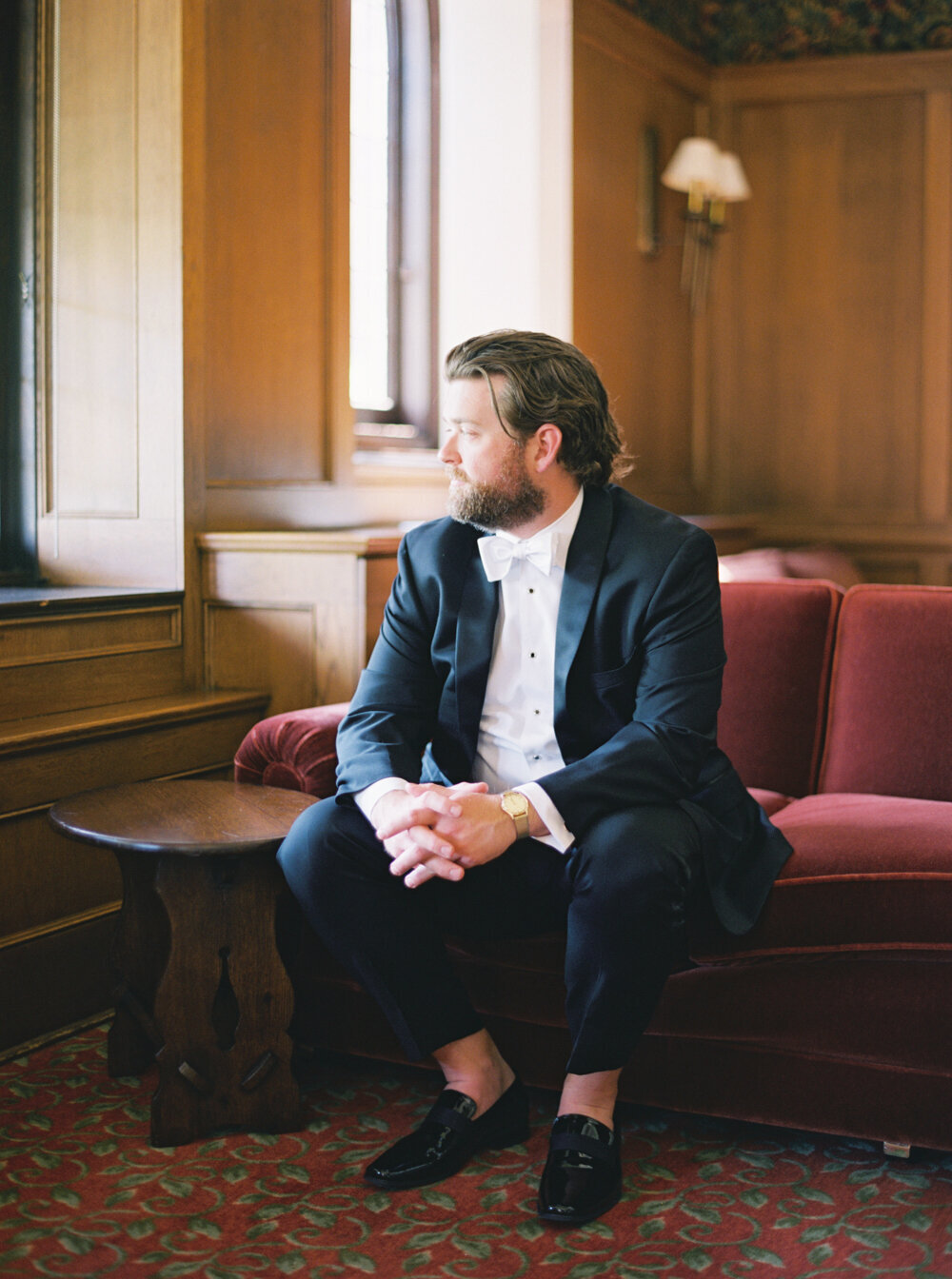 portrait of groom in black suit on red velvet couch in historic venue