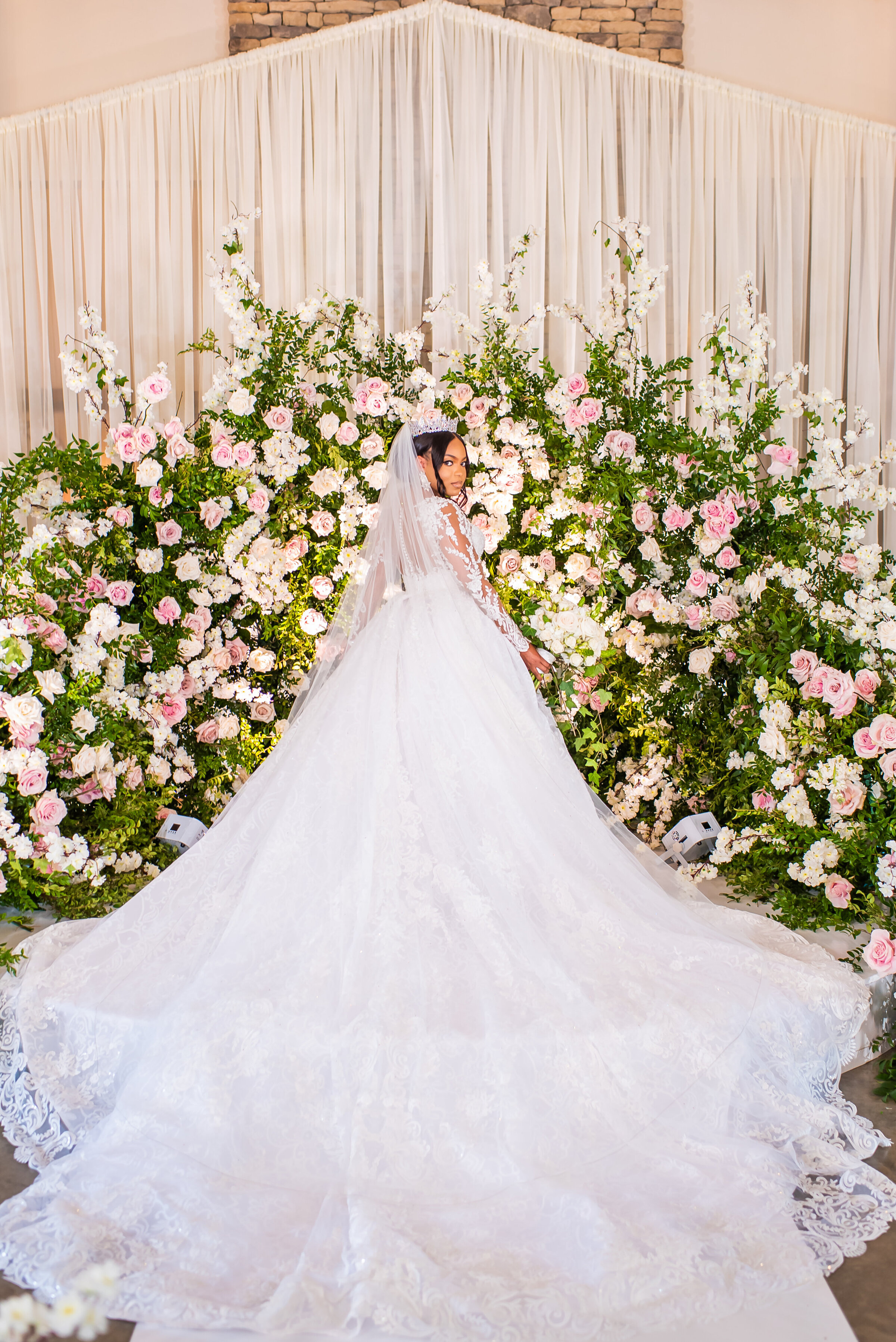 Bridal portrait in lace gown and tiara holding a white rose bouquet in front of lush floral wall at a romantic wedding ceremony in Birmingham, AL by Wells Design Co.