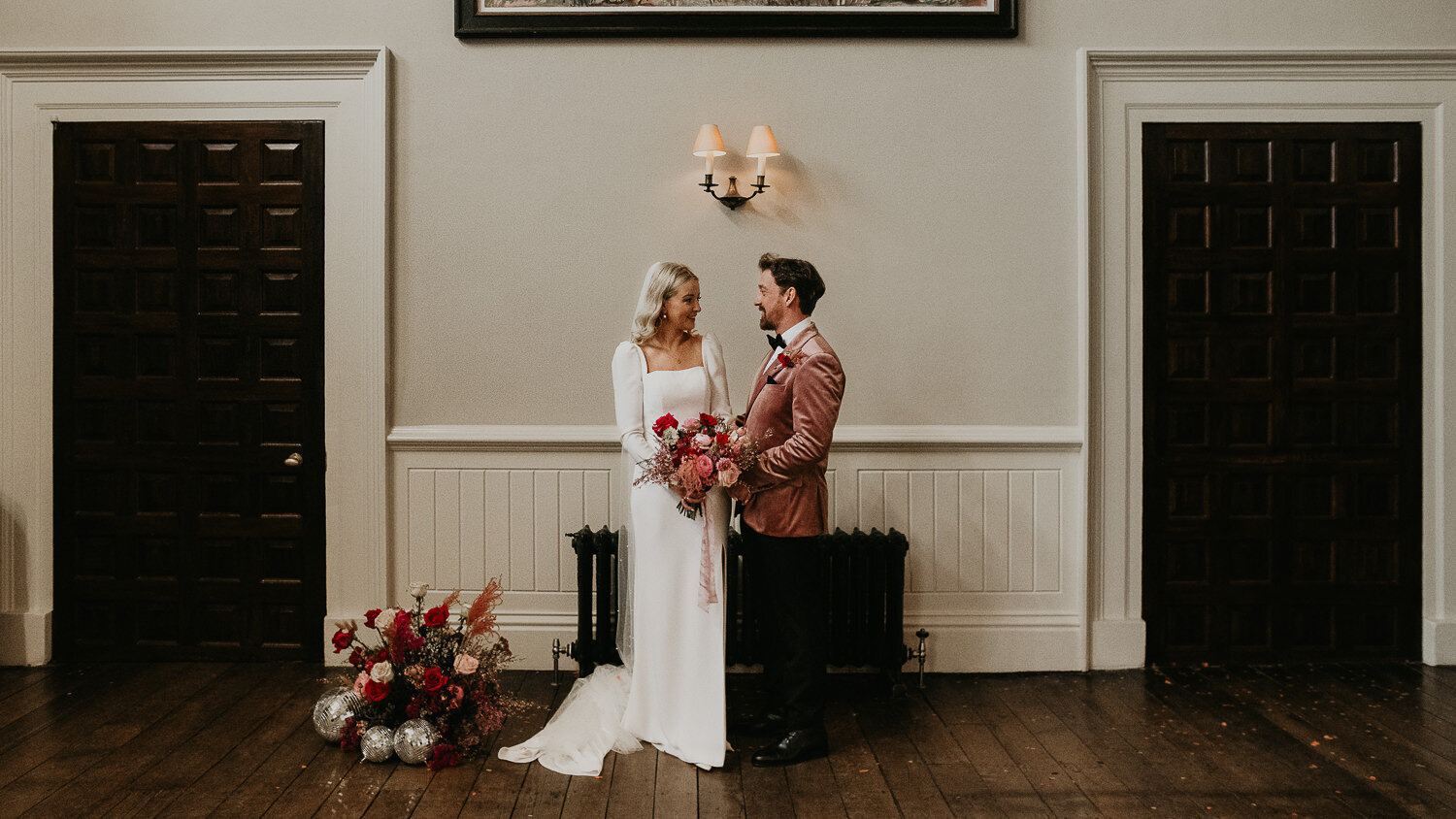 A wedding couple stand in the ceremony room of Elmore Court next to some pink wedding flowers.