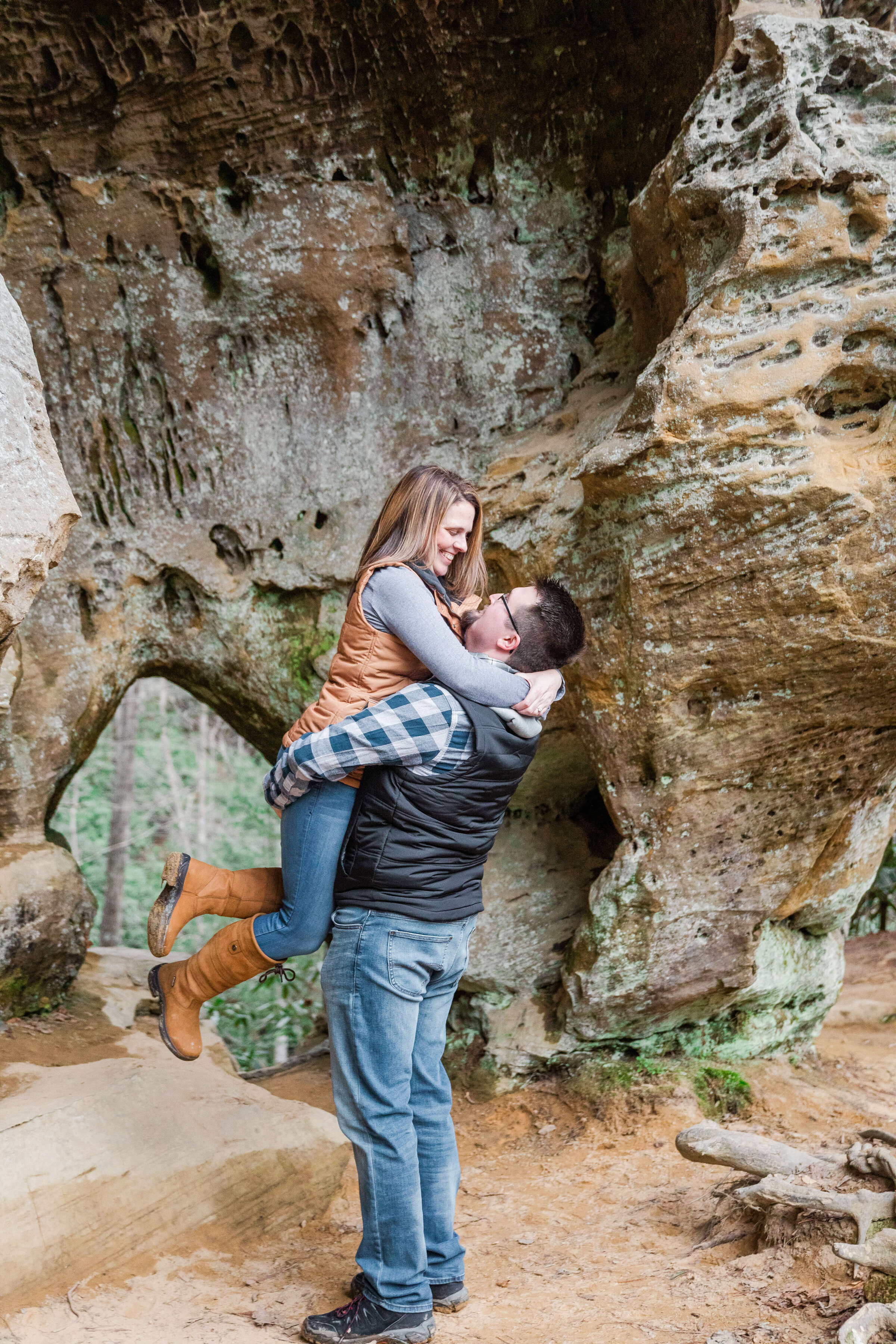 A man holds picks a woman up as they playfully laugh with one another. They are in a gorge on a hiking trail