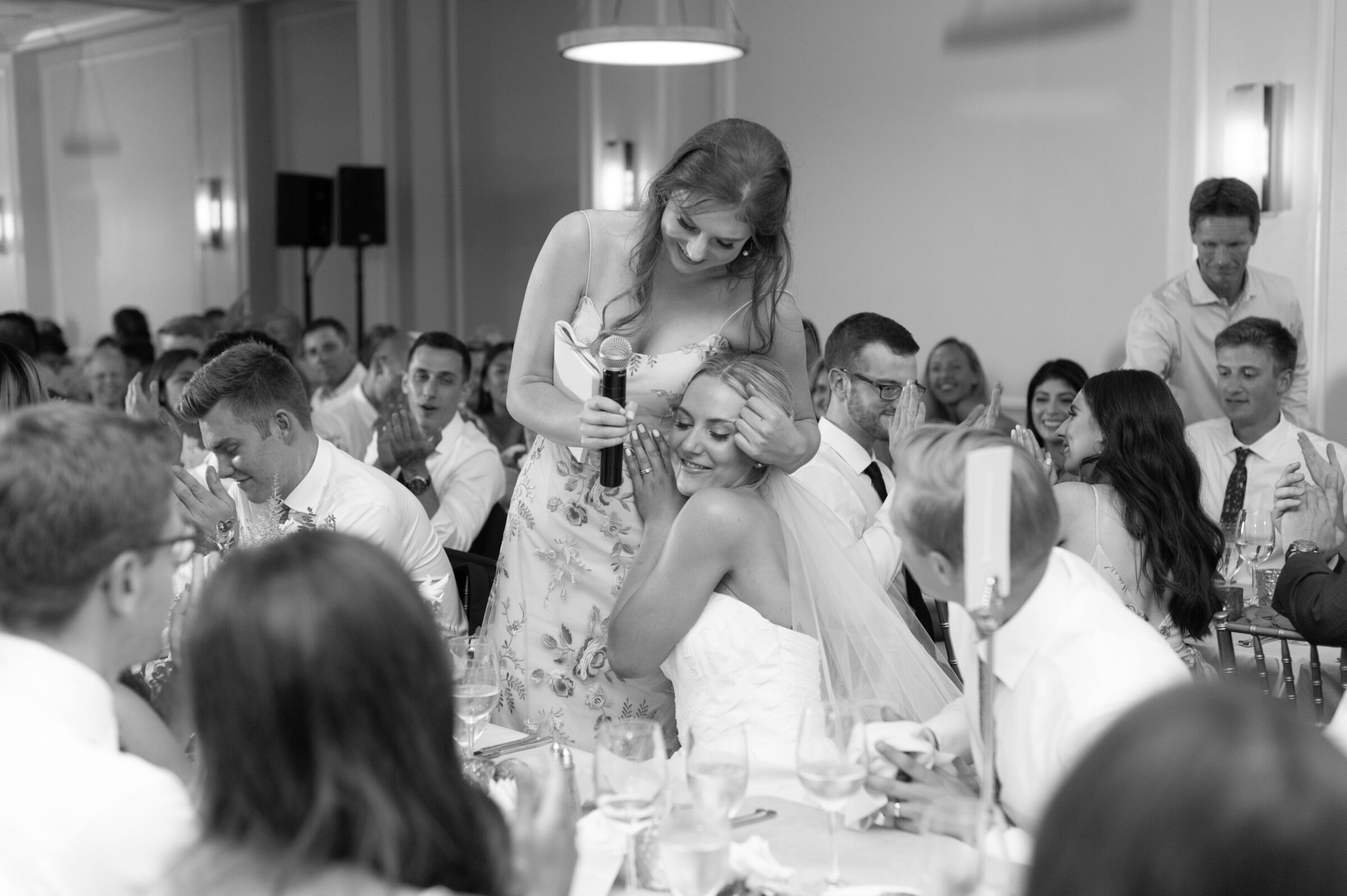 brides sister hugs her while she is sitting in black and white photo at wedding reception