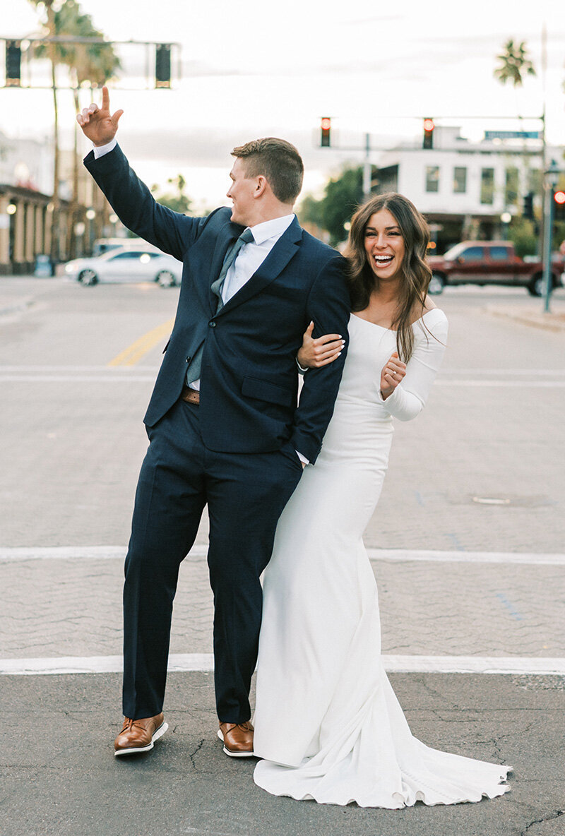 A newlywed couple celebrates in the middle of Arizona Avenue near SoHo63, a Chandler wedding and event venue. The groom raises his arm in excitement while the bride laughs, capturing the vibrant energy of a downtown Chandler wedding day.