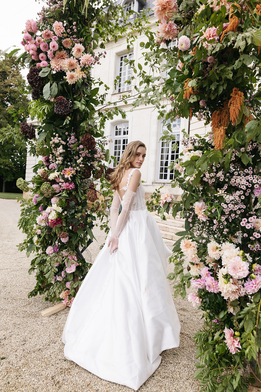 A bride in a white wedding gown walks through a vibrant floral arch featuring pink, white, and green blooms at a French chateau.
