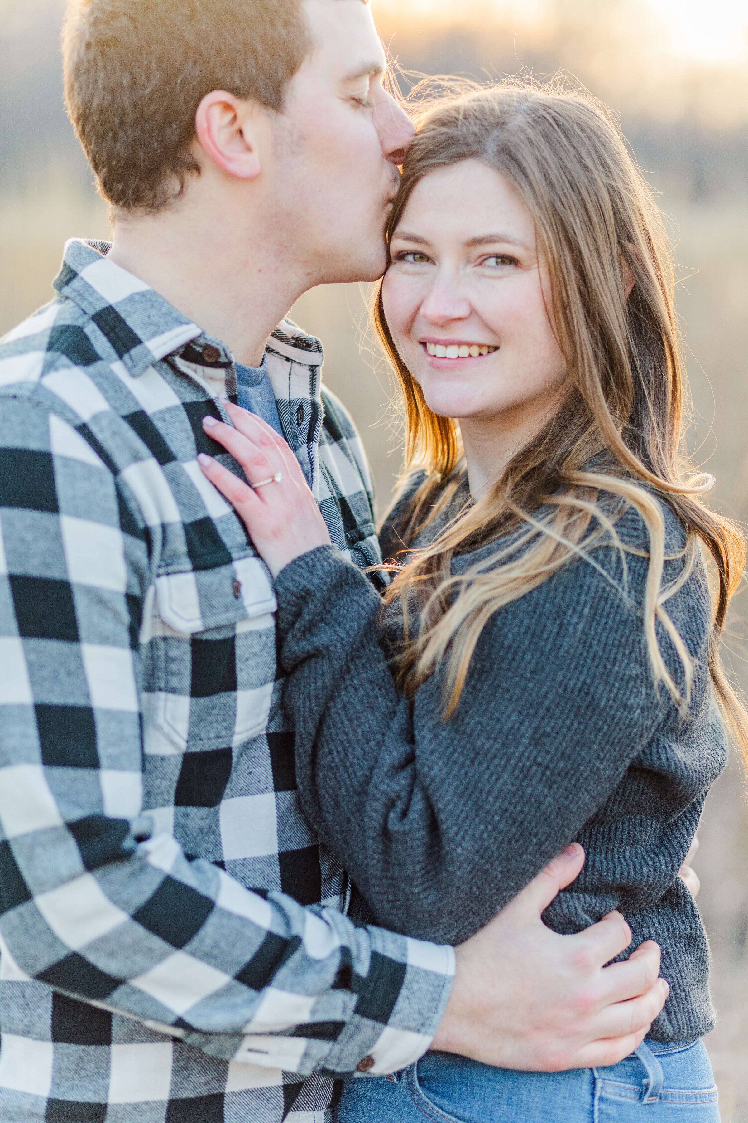 A man in a black and white plaid shirt holds his fiance who is wearing a grey sweater. She is smiling at the camera and he is kissing her temple. The sun is glowing in the back of them.