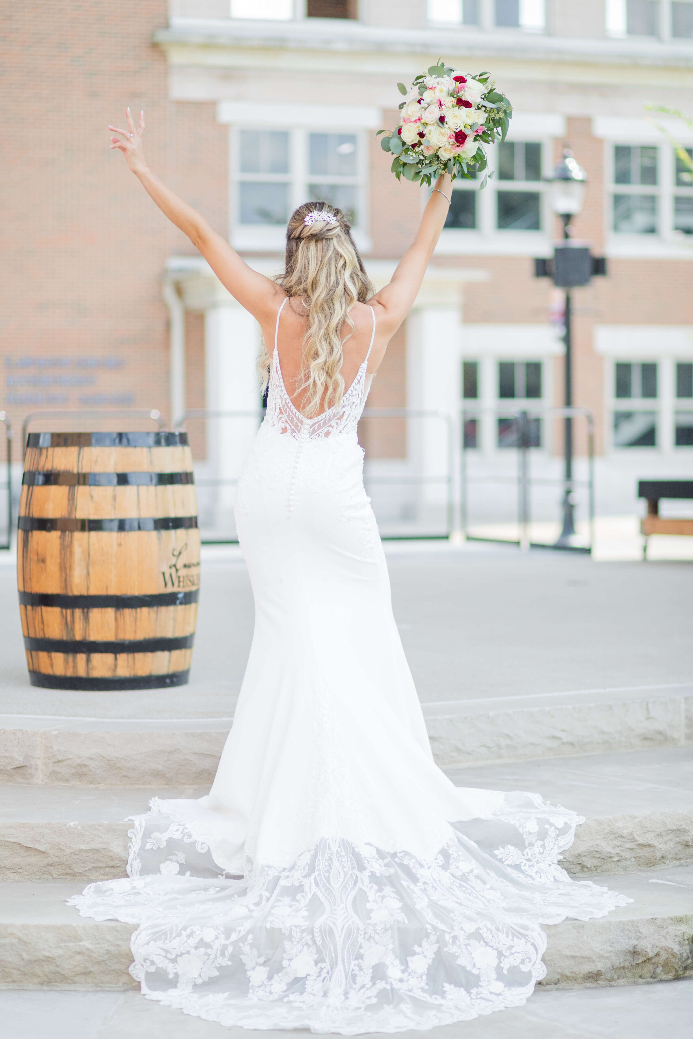 A bride has her back to the camera as she waves her hands in the area making the peace sign with one hand and holding her bouquet in the other.