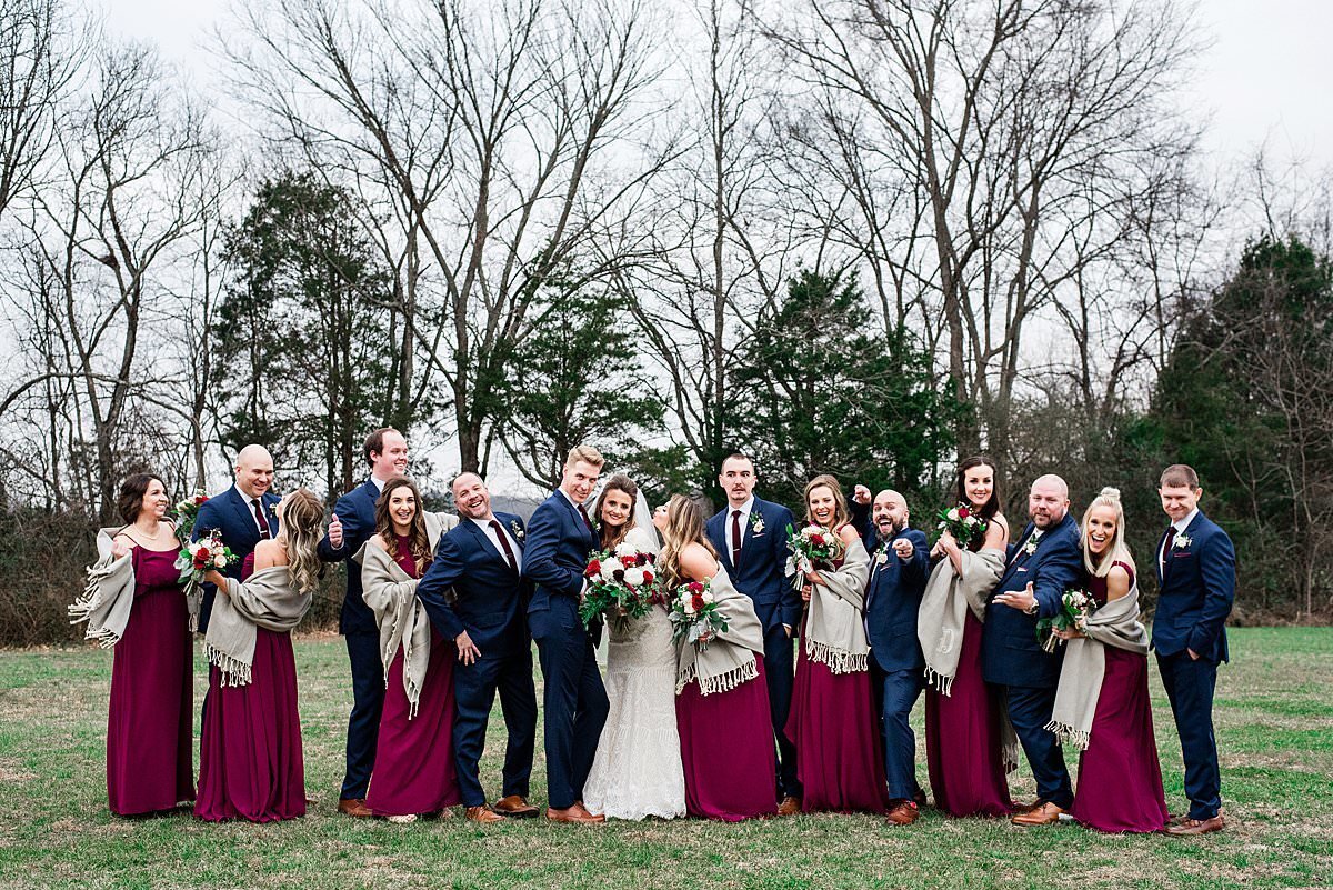 Full wedding party wearing burgundy and blue attire standing in a grass field at Grace Valley Farm during the winter, the girls have custom shawls