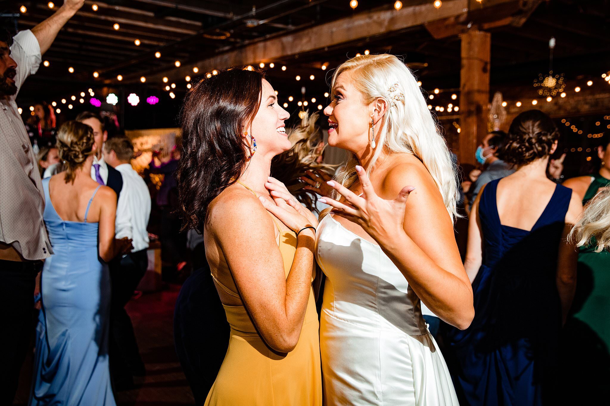 Bride and her friend dancing and singing facing one another on dance floor with bistro lights in the background of Marathon Village
