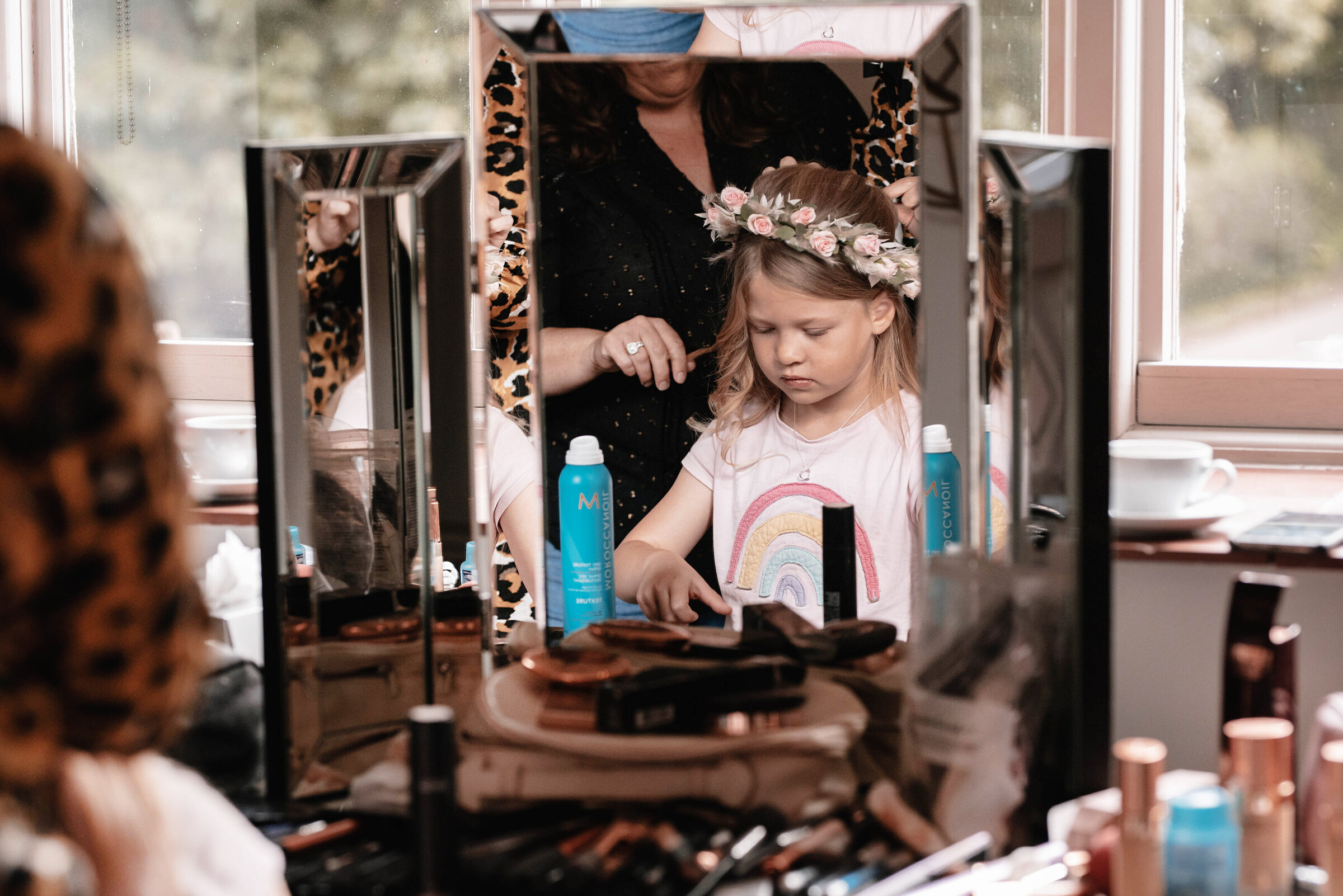 Reflection of a young flower girl having her hair styled with floral headband surrounded by make-up and hair products