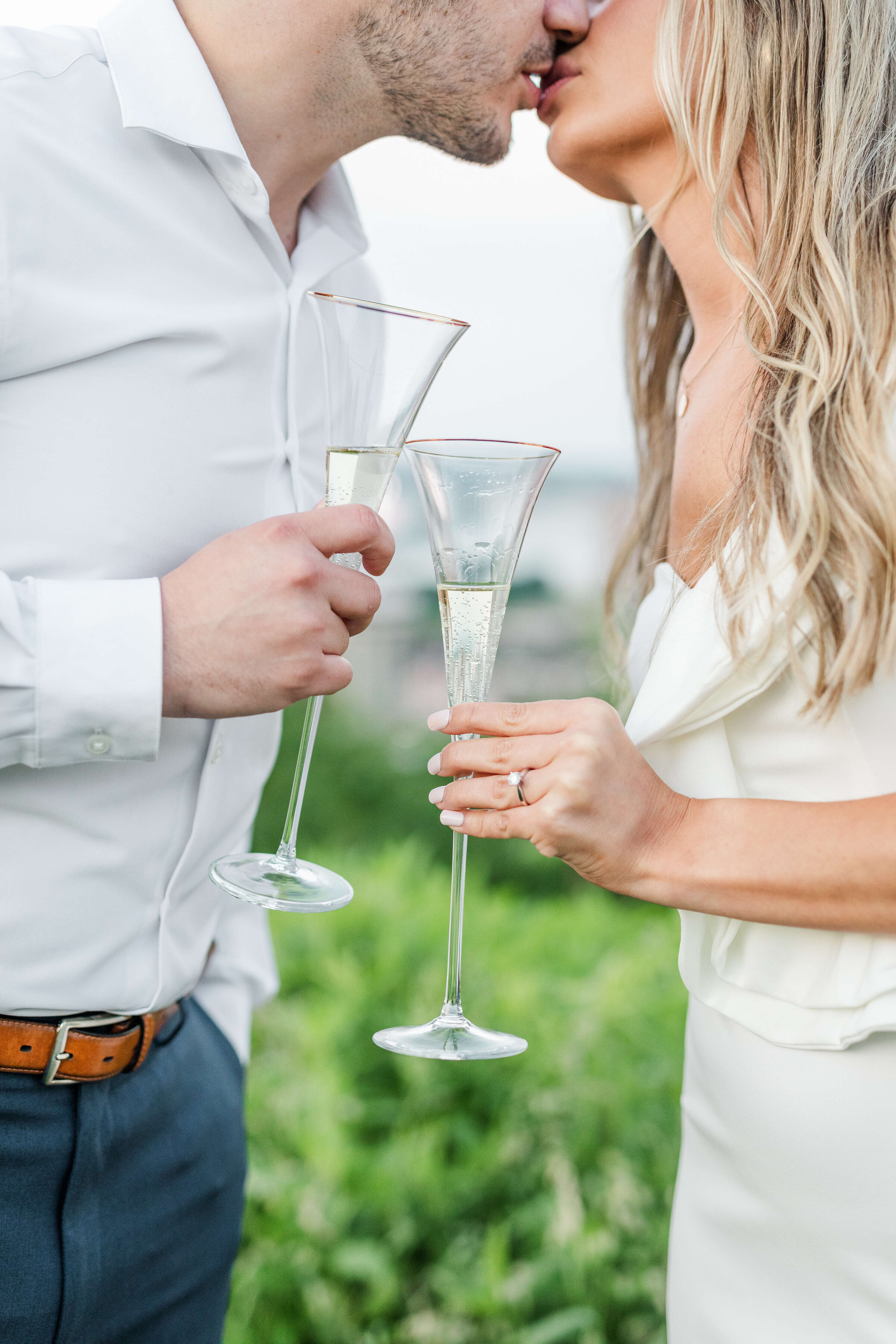 A man and woman kiss as they hold their champagne glasses together. They are celebrating their engagement