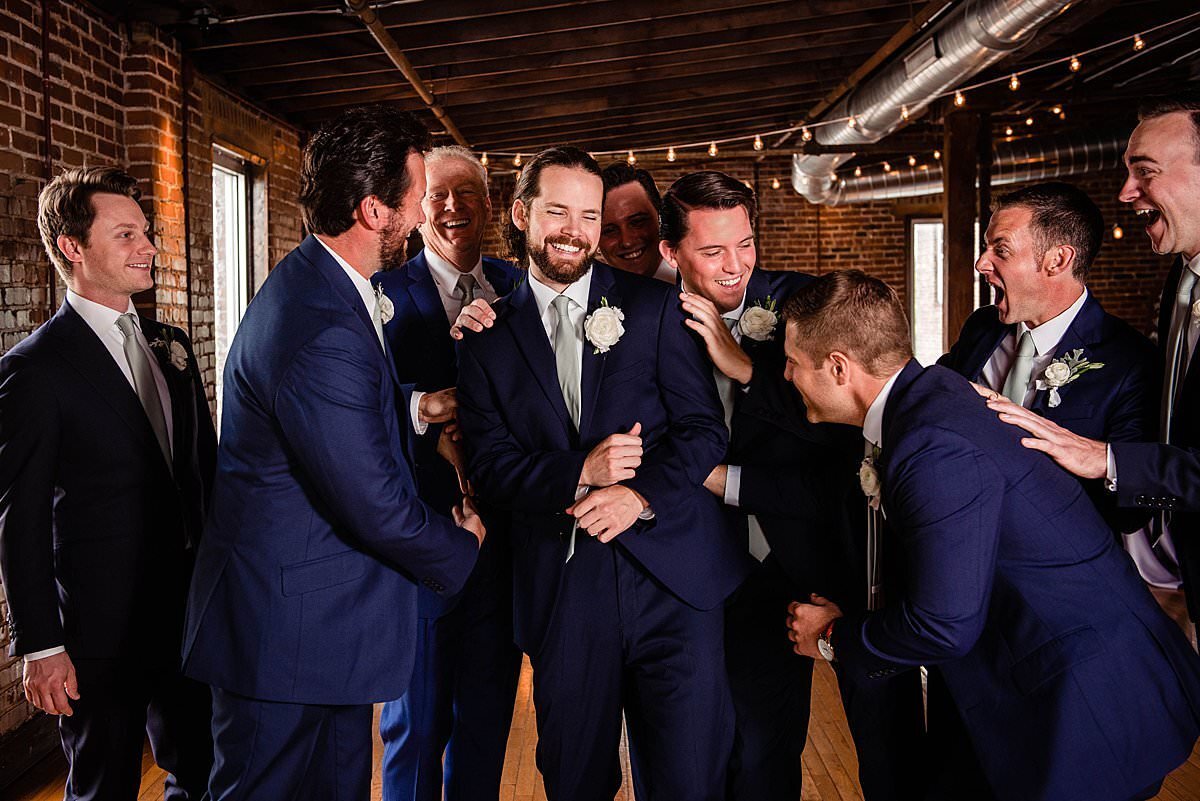 Groomsmen laughing with groom  during group photos in the bar at Cannery ONE Nashville, the men are wearing navy suits and bistro lights can be seen in the background