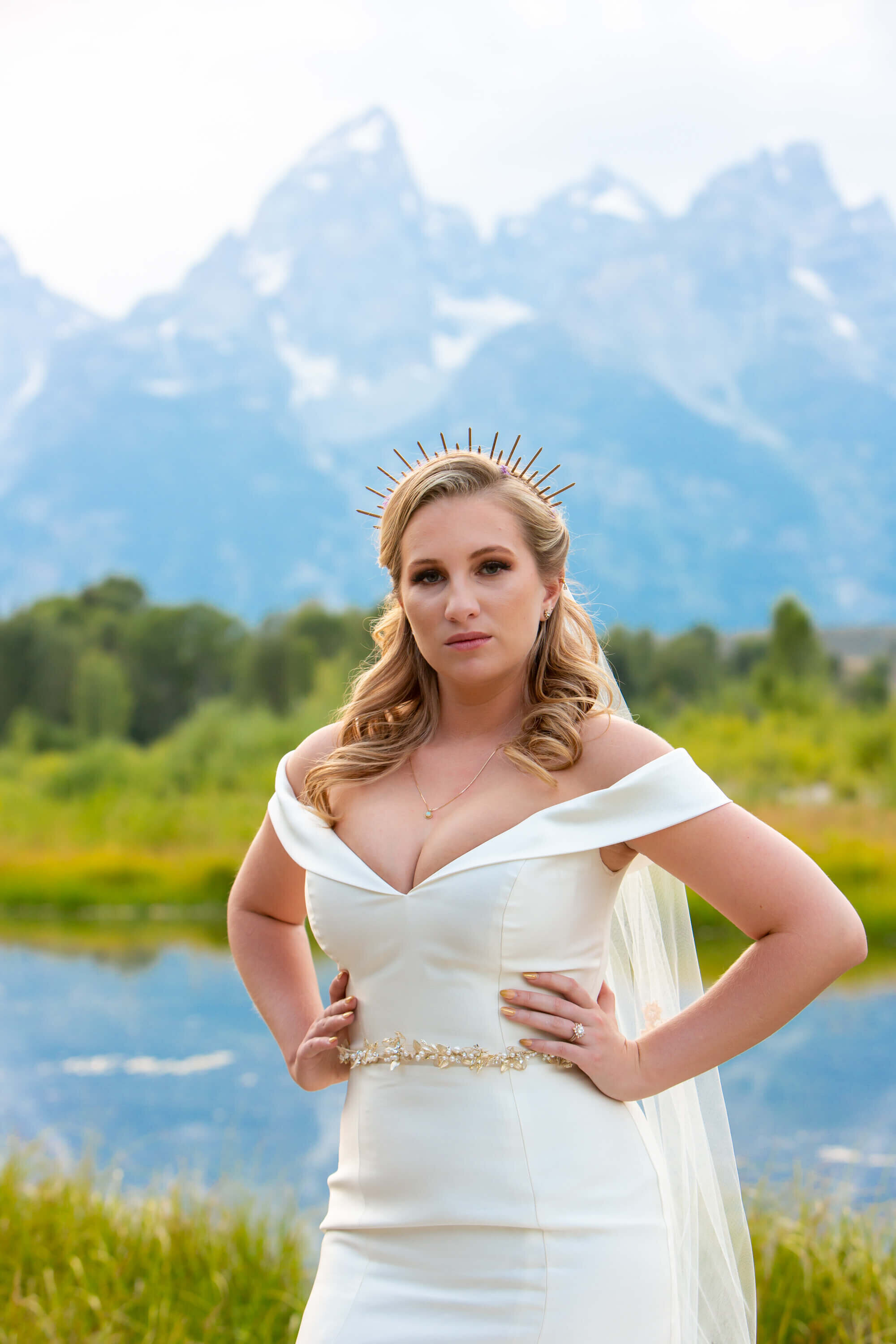 A bride stands confidently in front of the Teton mountains, wearing an off-the-shoulder satin wedding gown and a gold headpiece.