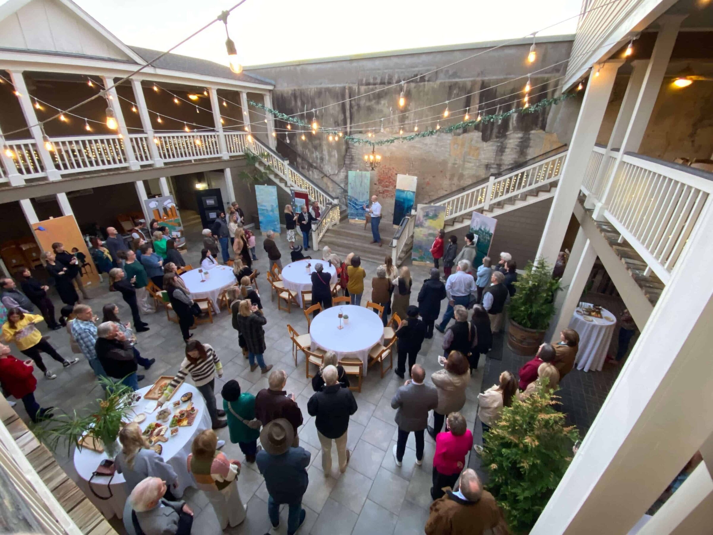 A man stands on stairs speaking to a gathered crowd at an art exhibit on the stairs in an open courtyard. Large paintings are displayed around him, and people listen and observe from below.