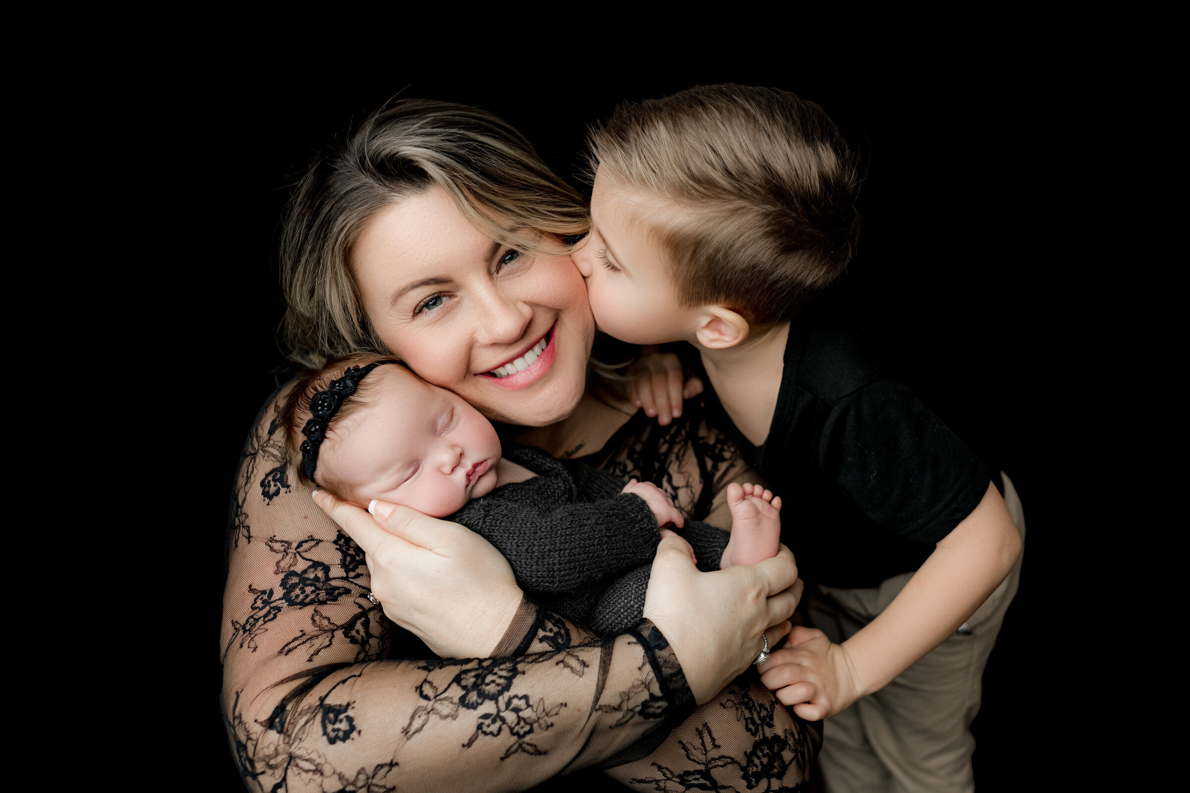 A smiling woman holds a sleeping baby in her arms as a young boy kisses her cheek. She wears a lace top, the baby is in a black outfit with a headband, and the boy is dressed in a black shirt. This touching moment is beautifully captured by a North Carolina newborn photographer against a plain black background.