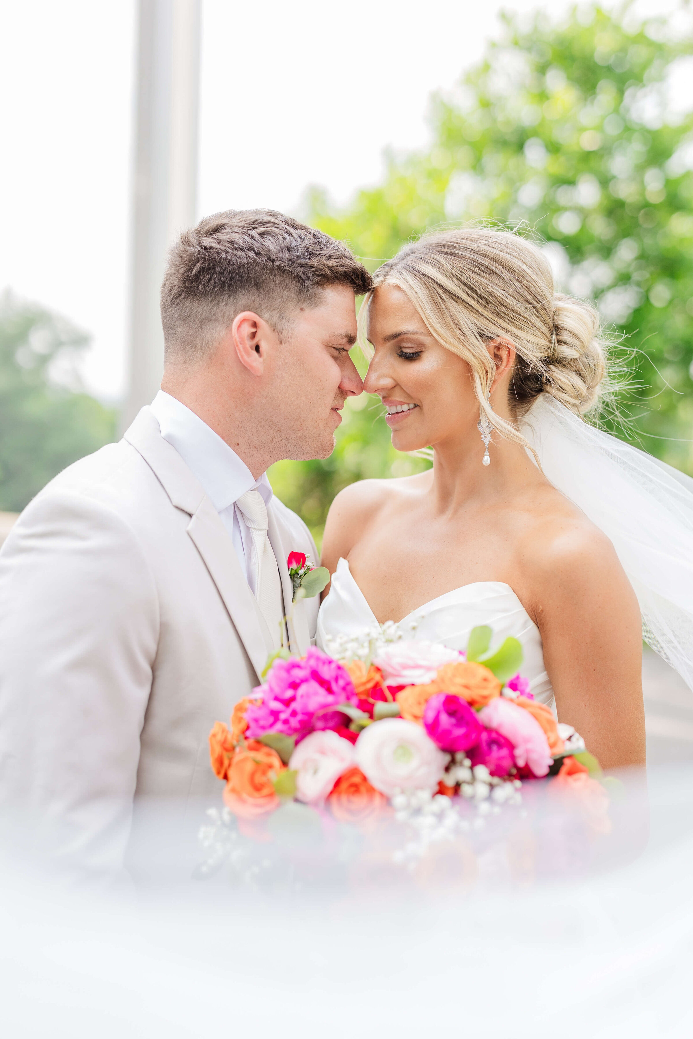 A bride and groom stand facing each other smiling. The groom is wearing a tan suit and the bride is holding a bright pink bouquet. It's summer, and the trees are bright green in the back of them.