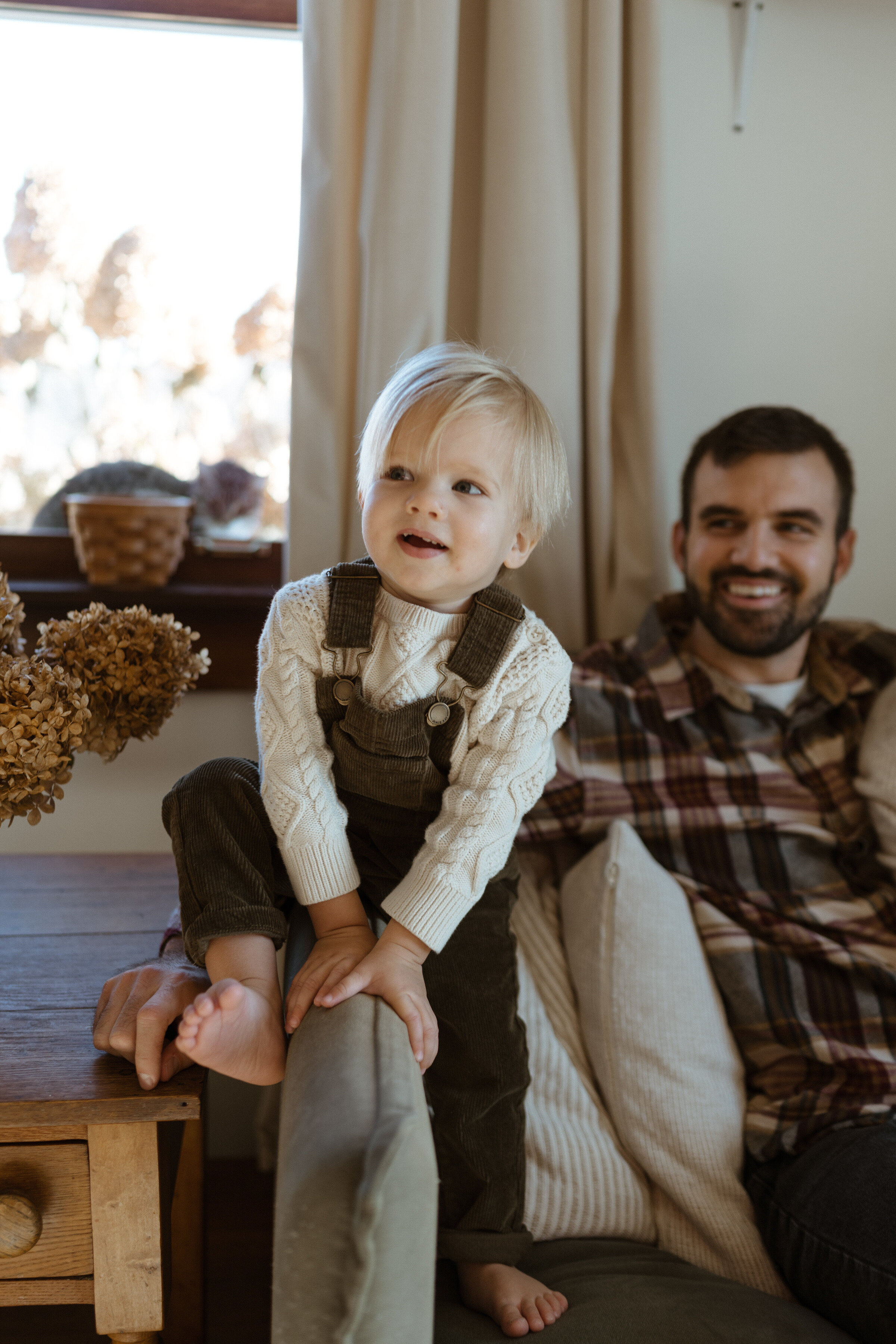 A young blond toddler  boy in green overalls sits on the arm of a couch smiling