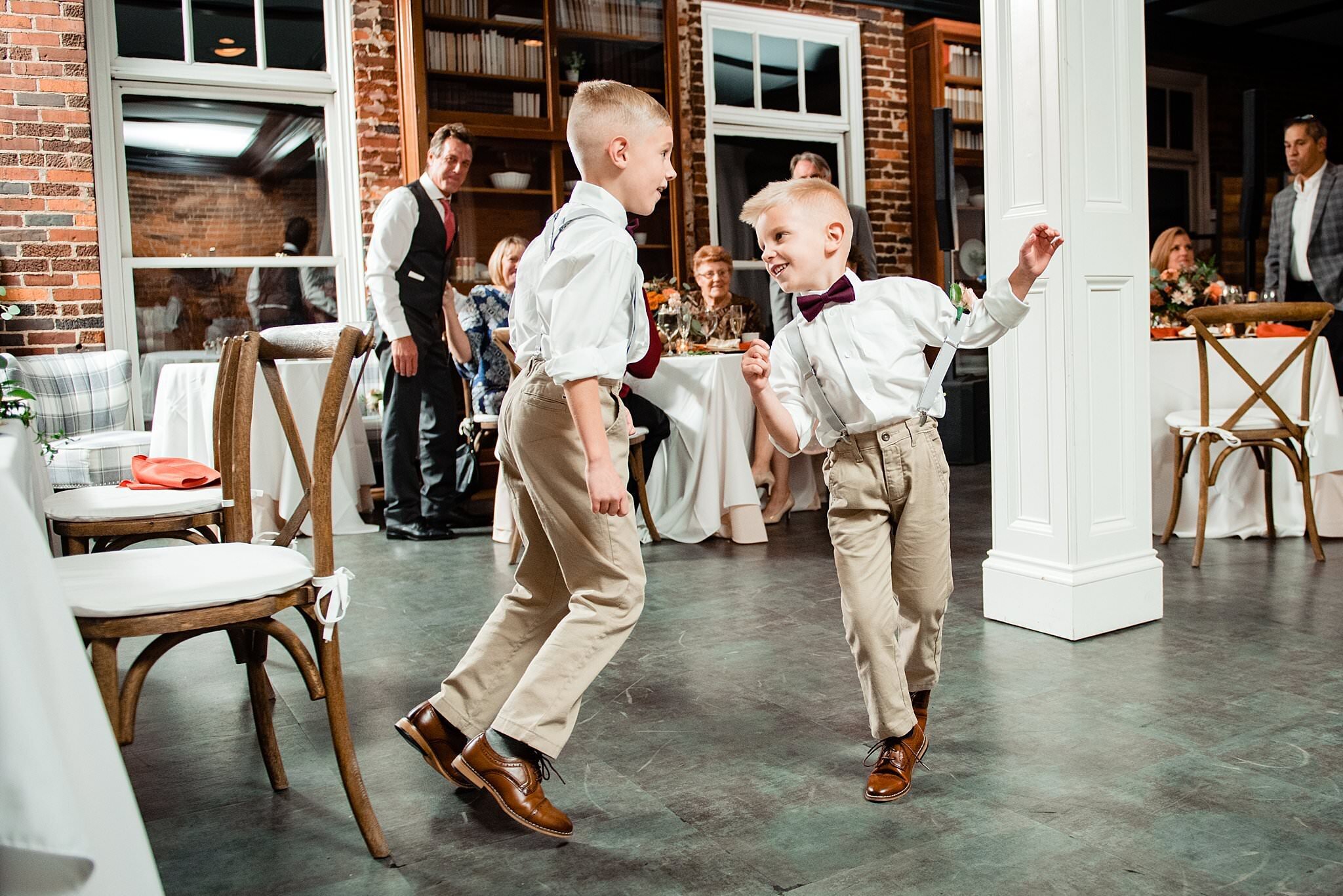 Ring bearers wearing suspenders and bow ties dancing in circles together on the dance floor at the event space at Tasty Table