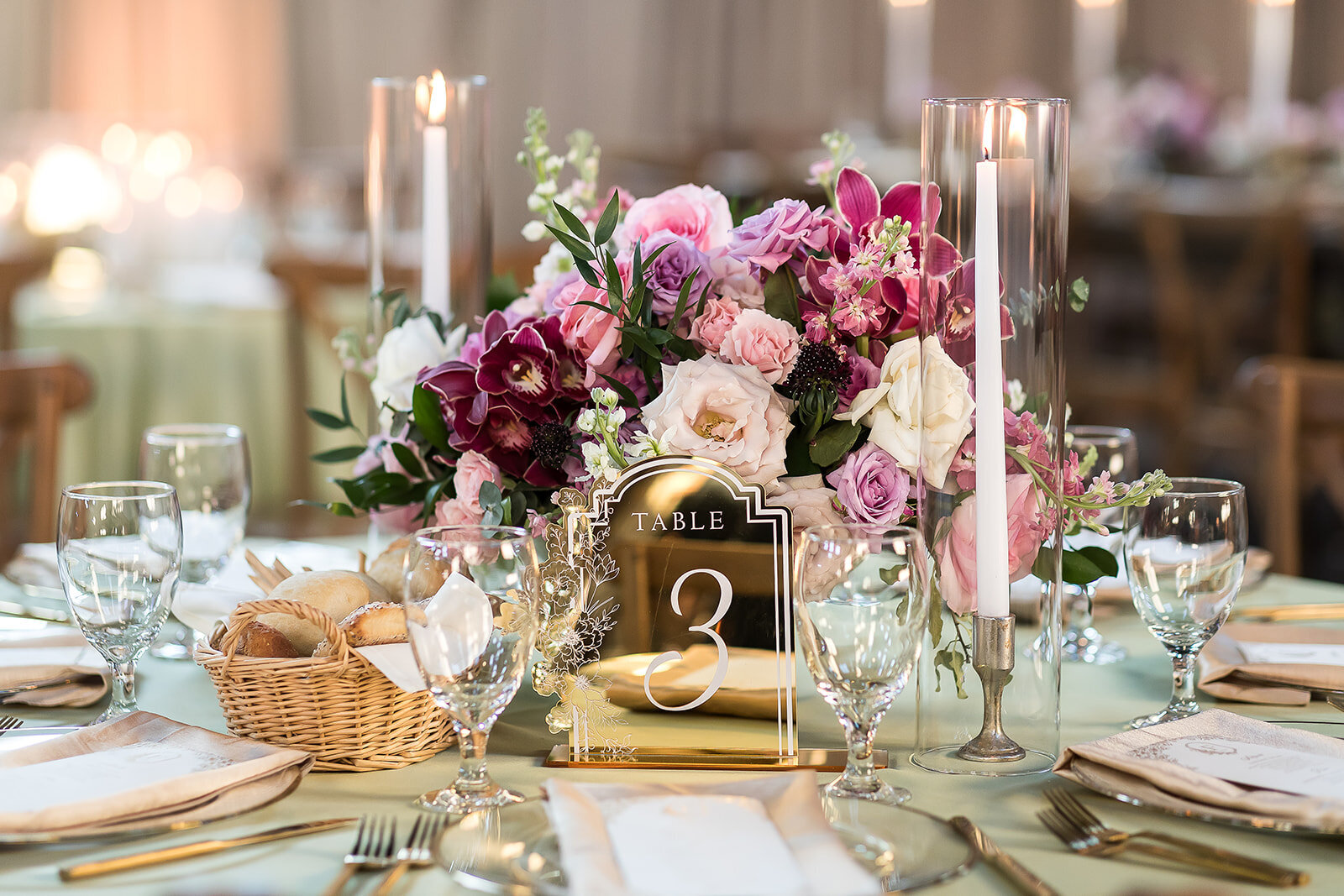 Elegant guest table with soft green linen, pink and mauve floral centerpiece, tapered candlelight, and gold mirrored table number at The Kent Memphis wedding reception by Wells Design Co.