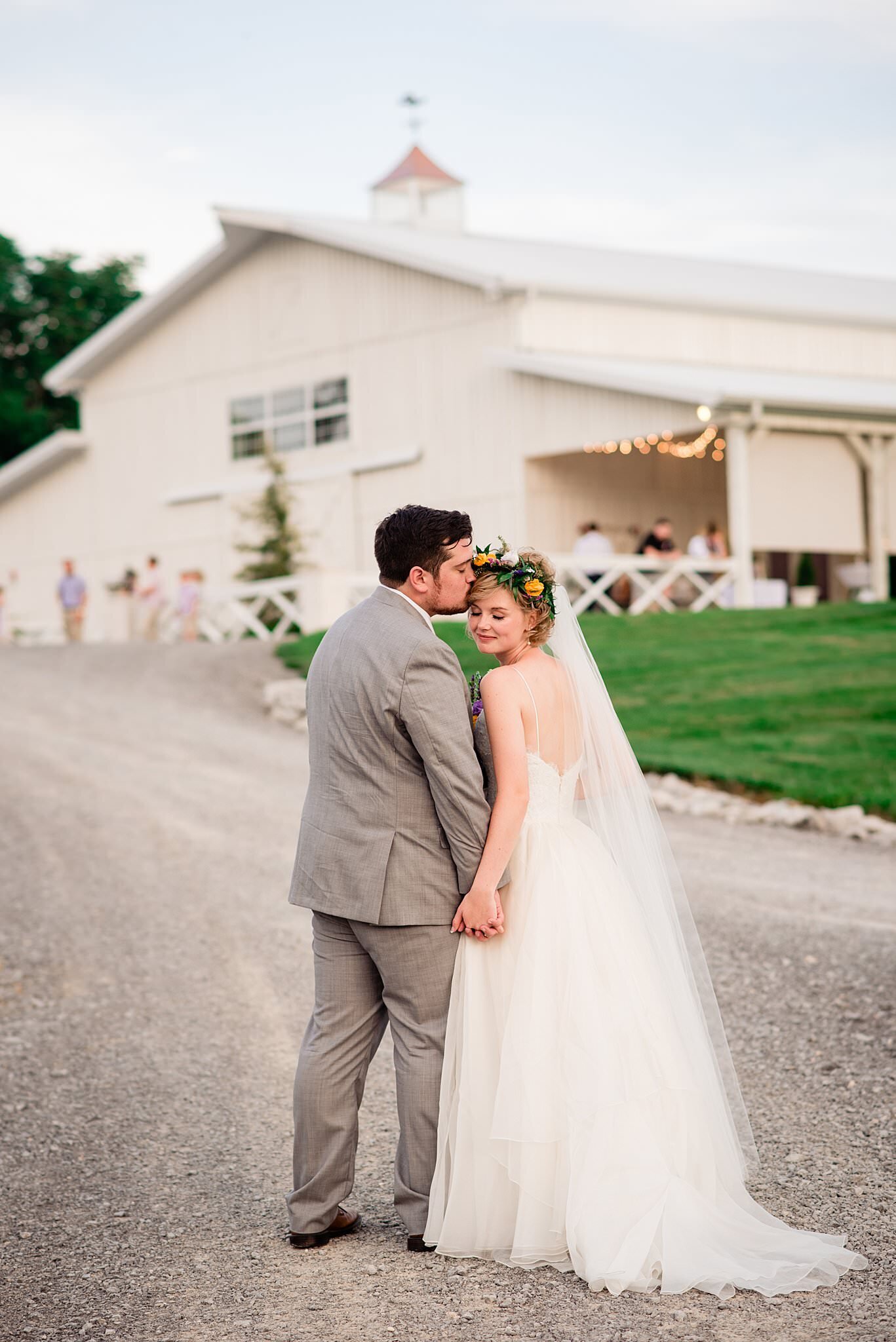 Bride and groom holding hands standing on gravel path with their backs to the camera as he kisses her temple. She has a long veil on and a flower crown and White Dove Barn is in the background