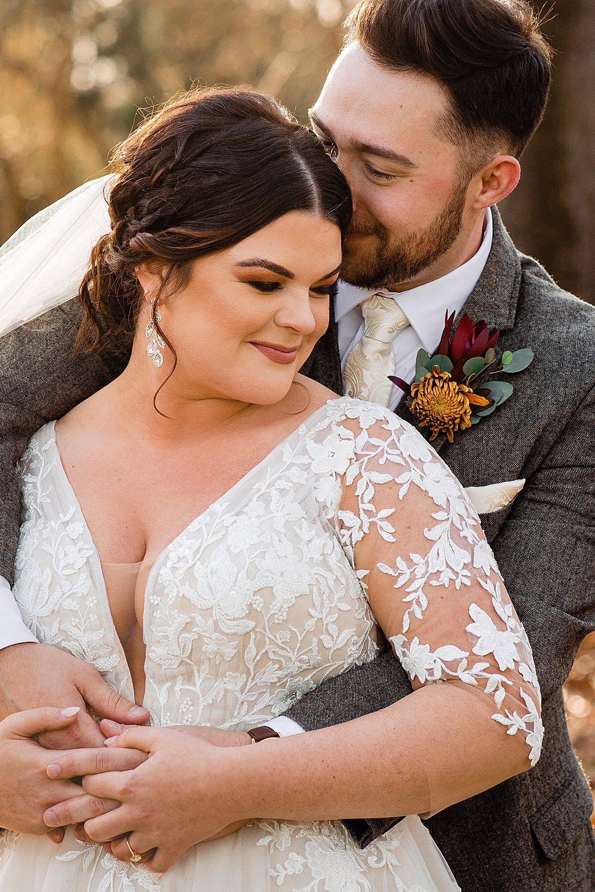 Bride wearing long sleeve lace dress and groom with a grey tweed suit holding his wife who has braids in her updo