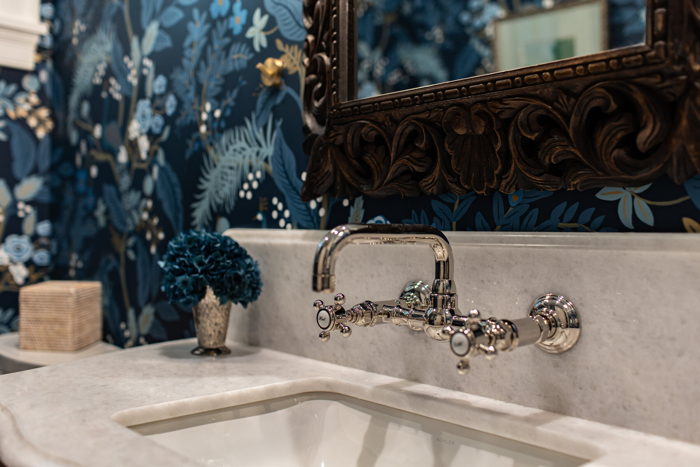A detailed close-up of a bathroom sink featuring a marble countertop, silver fixtures, and vibrant floral blue wallpaper. The combination of luxury materials and bold design creates an elegant and eye-catching focal point in the space.