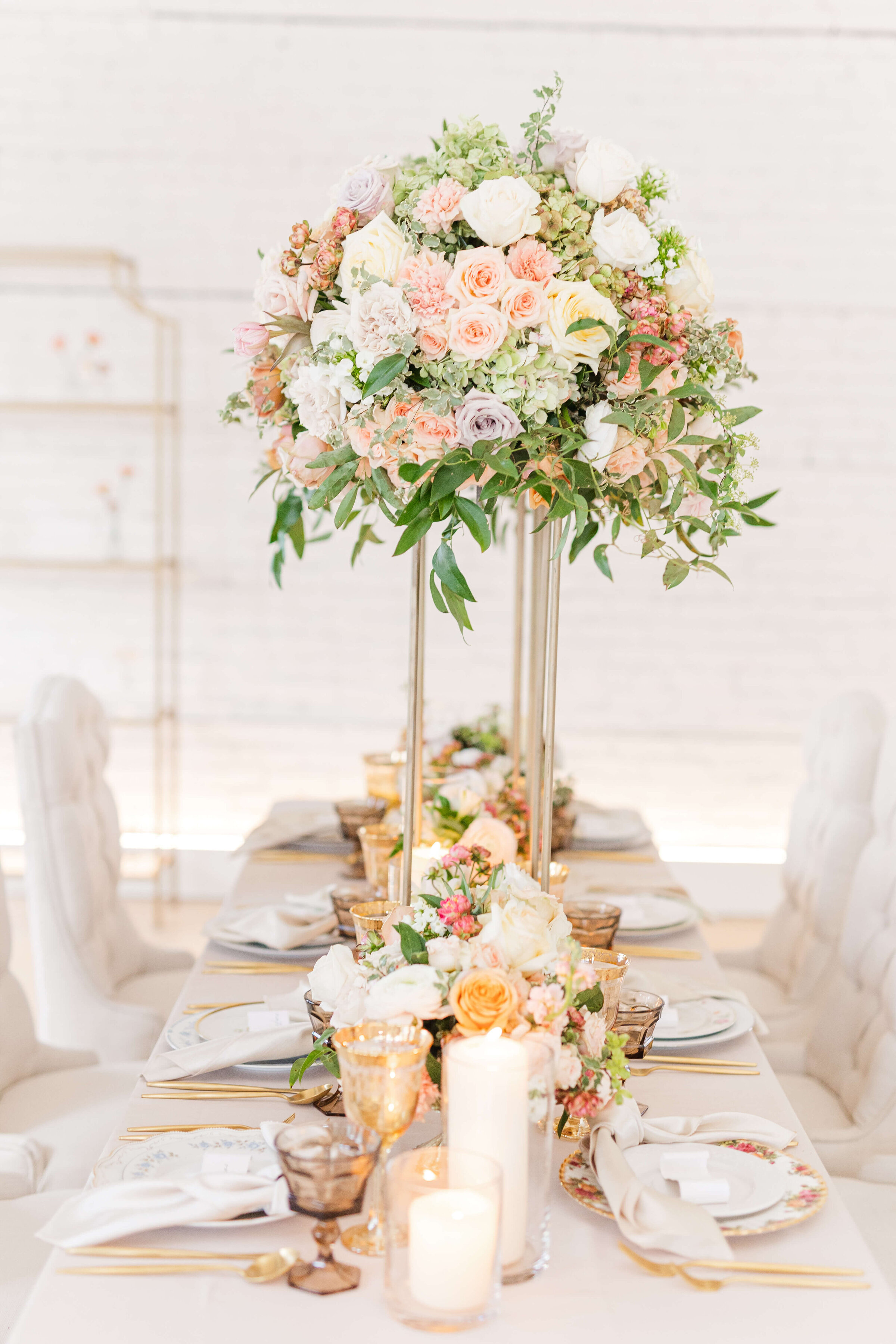 A wedding reception table with a tall floral centerpiece full of pink, blush, and peach flowers. There are other flowers and rose glasses on the table. The table is ivory and so are the chairs