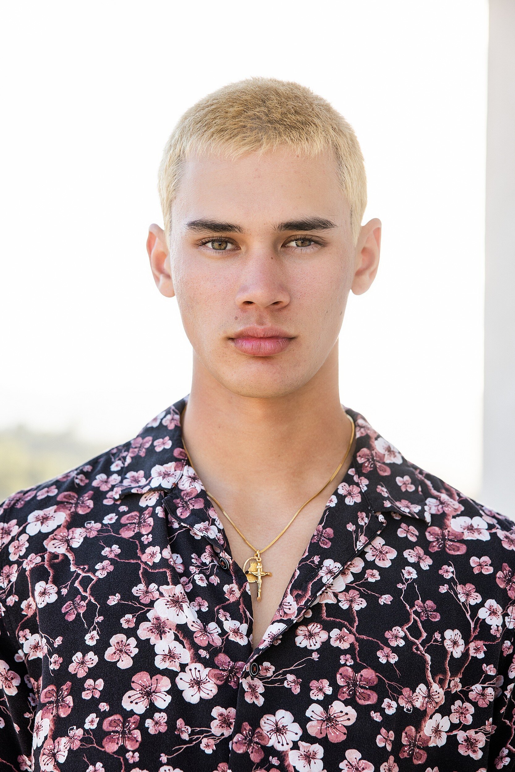 Striking headshot of a young man with bleached blonde hair and a serious expression, wearing a black button-up shirt with pink cherry blossom print and a gold cross necklace.

