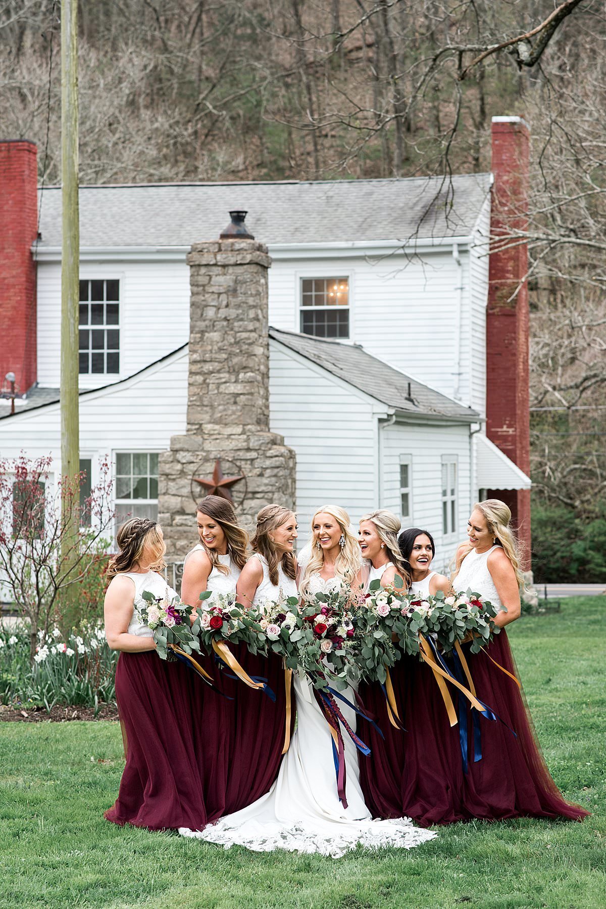 Bride grouped with bridesmaids who are wearing burgundy tulle skirts with white lace tops by historic home at Drakewood Farms