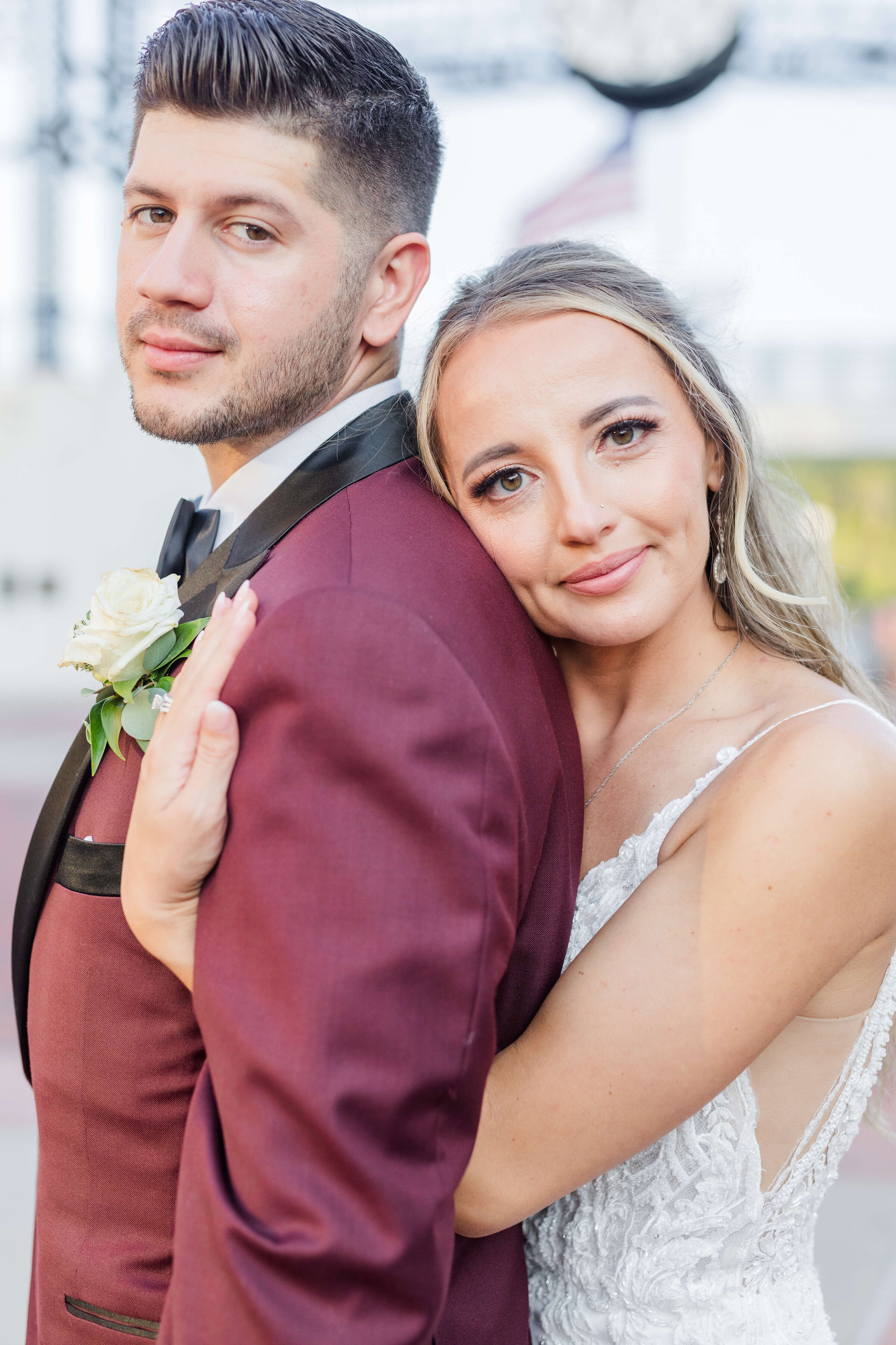 A groom in a maroon tux, looks at the camera as his bride hugs him from behind and she's also looking at the camera. The sun is glowing on them.