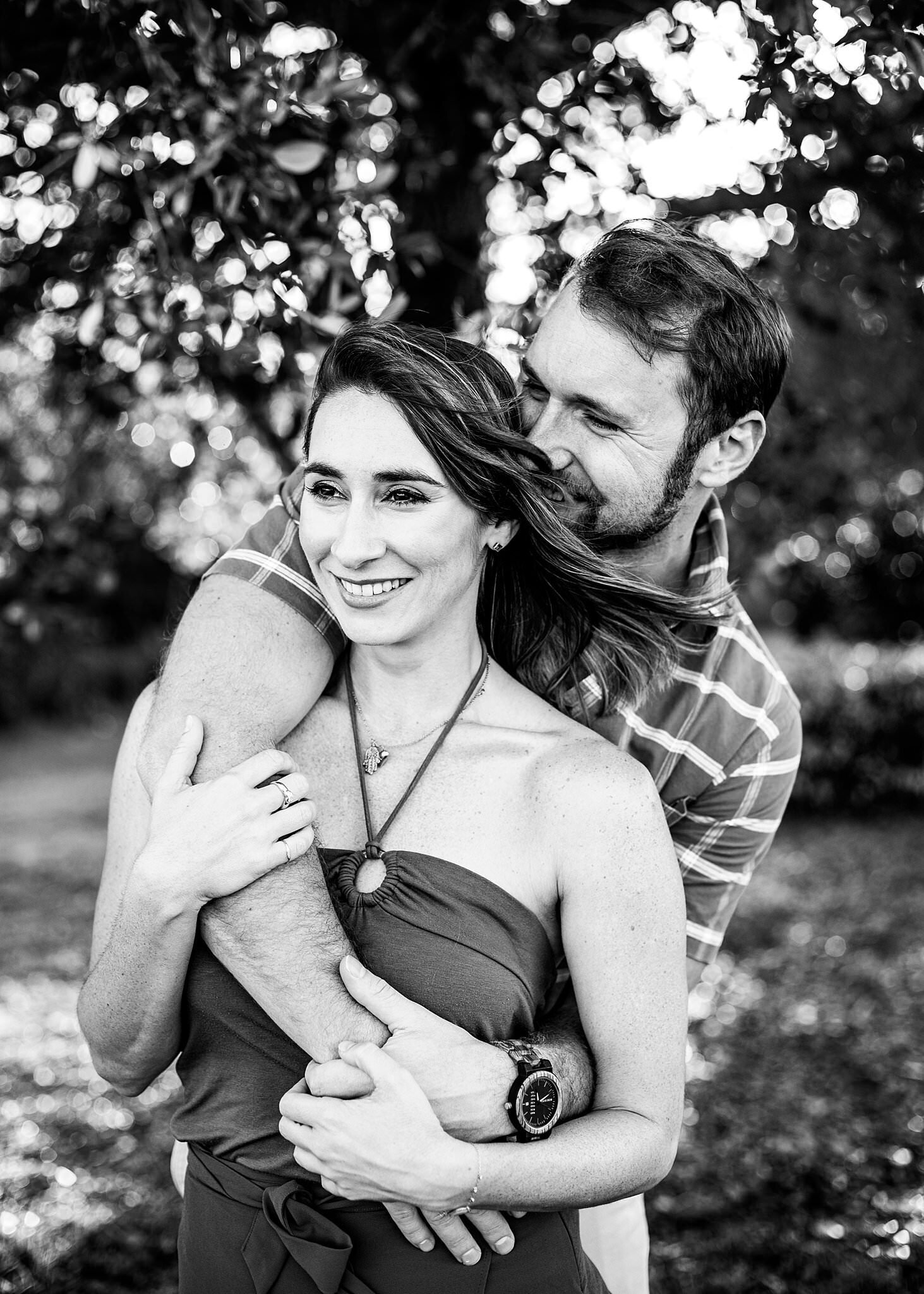 Black and white photo of couple, with man hugging her from the back and smiling