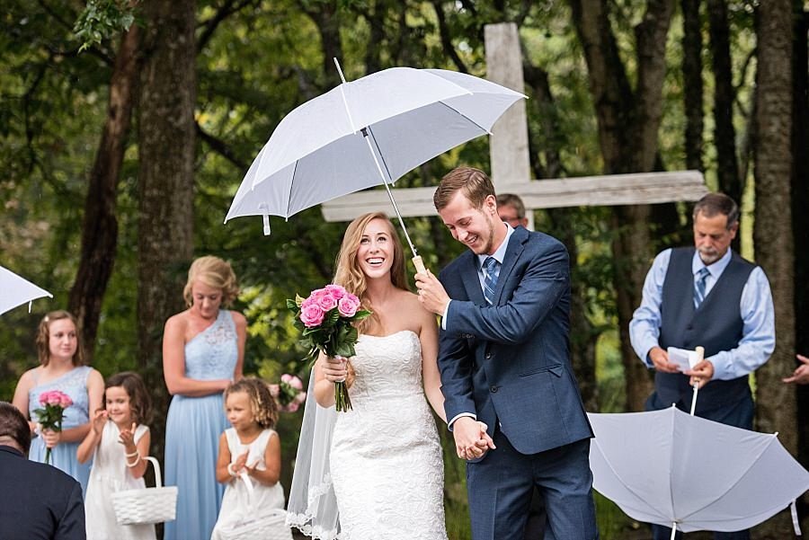 Groom holding white umbrella over him and his wife during outdoor rainy ceremony