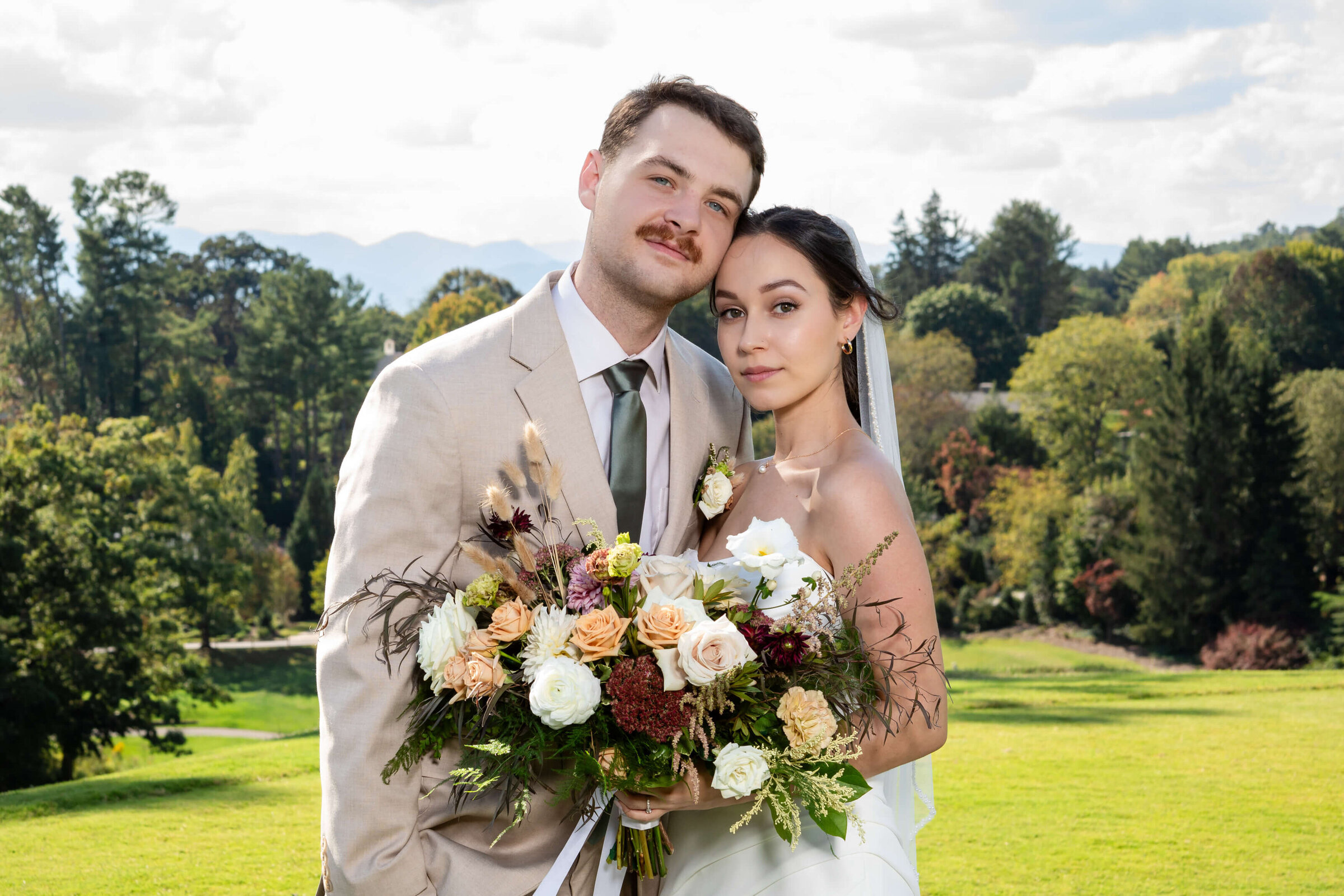 Modern bride in a sleek white gown and groom in a tan suit posing with a lush neutral‐toned bouquet, framed by grand mountain vistas near Asheville, North Carolina for a luxury wedding experience.
