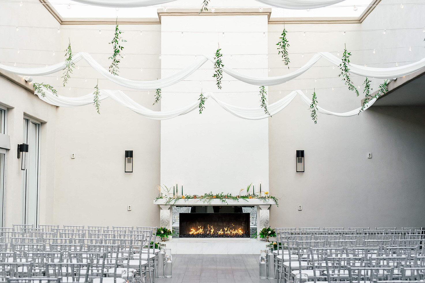 A bright and modern ceremony setup at SoHo63, a Chandler wedding and event venue, features rows of silver chairs facing a white fireplace framed with greenery and florals. Soft white drapery and hanging vines create a sophisticated and airy backdrop for a stylish Arizona wedding.