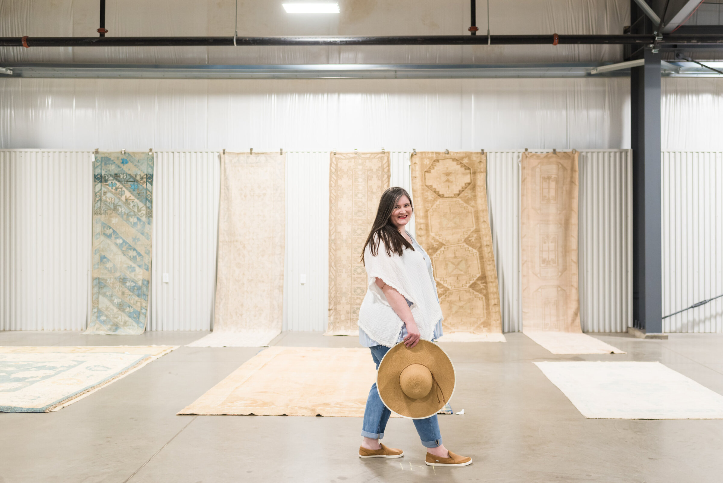 Lauren strolls past antique rugs at antique market while holding sun hat