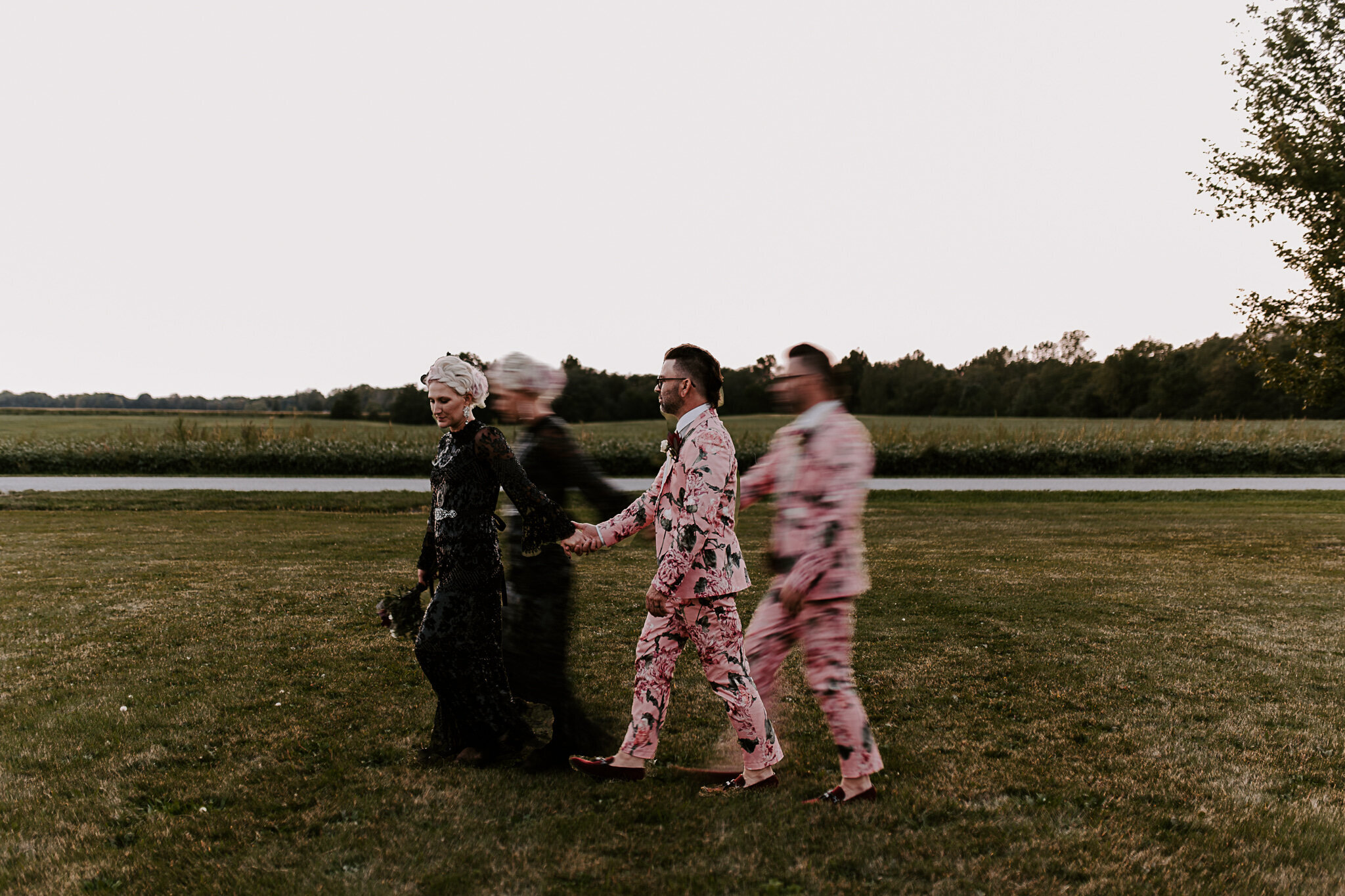 A bride and groom in a floral suit with tattooswalk across a field at sunset