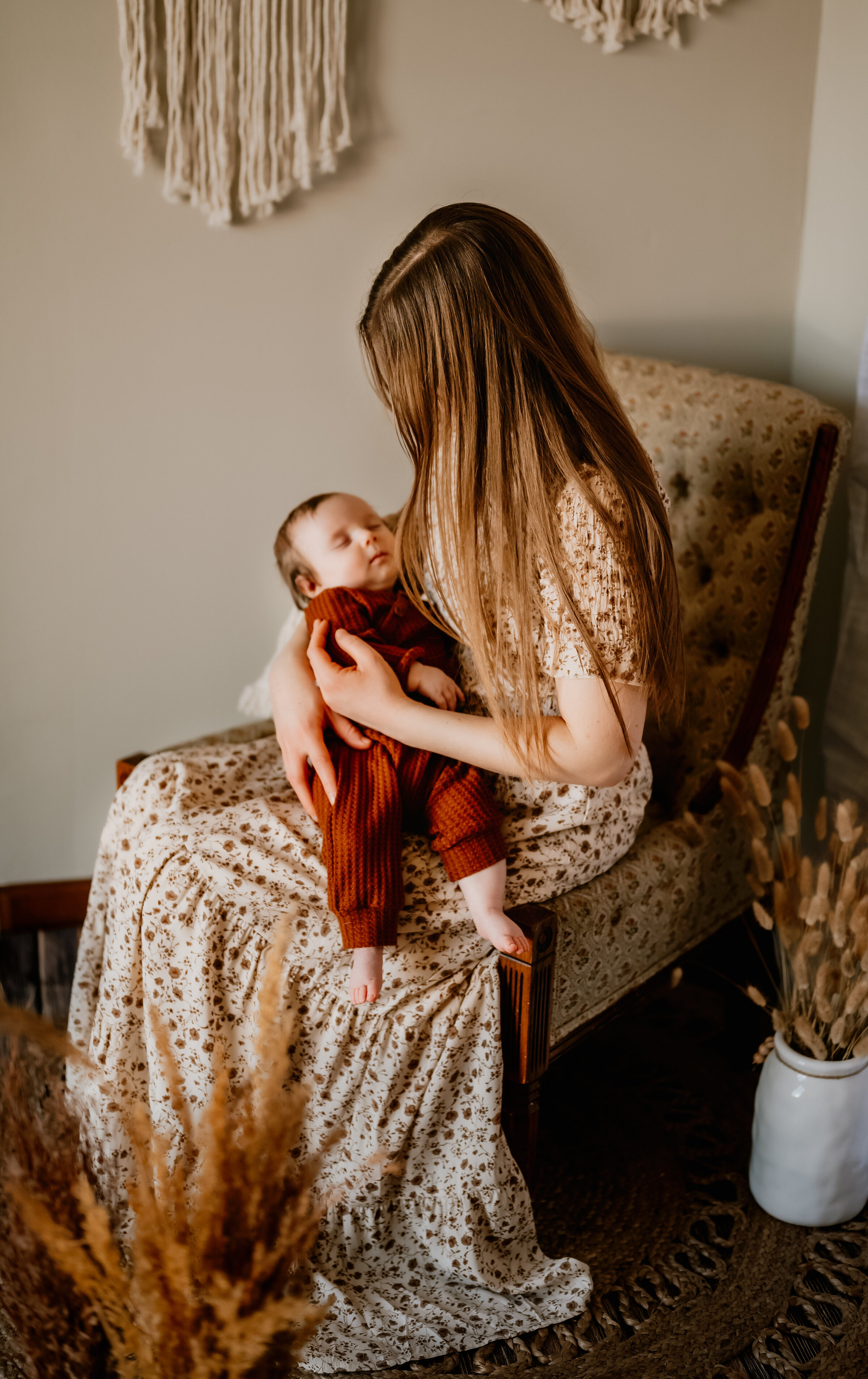 Mother holds her three month old in vintage wingback chair surrounded by dried grass in boho studio