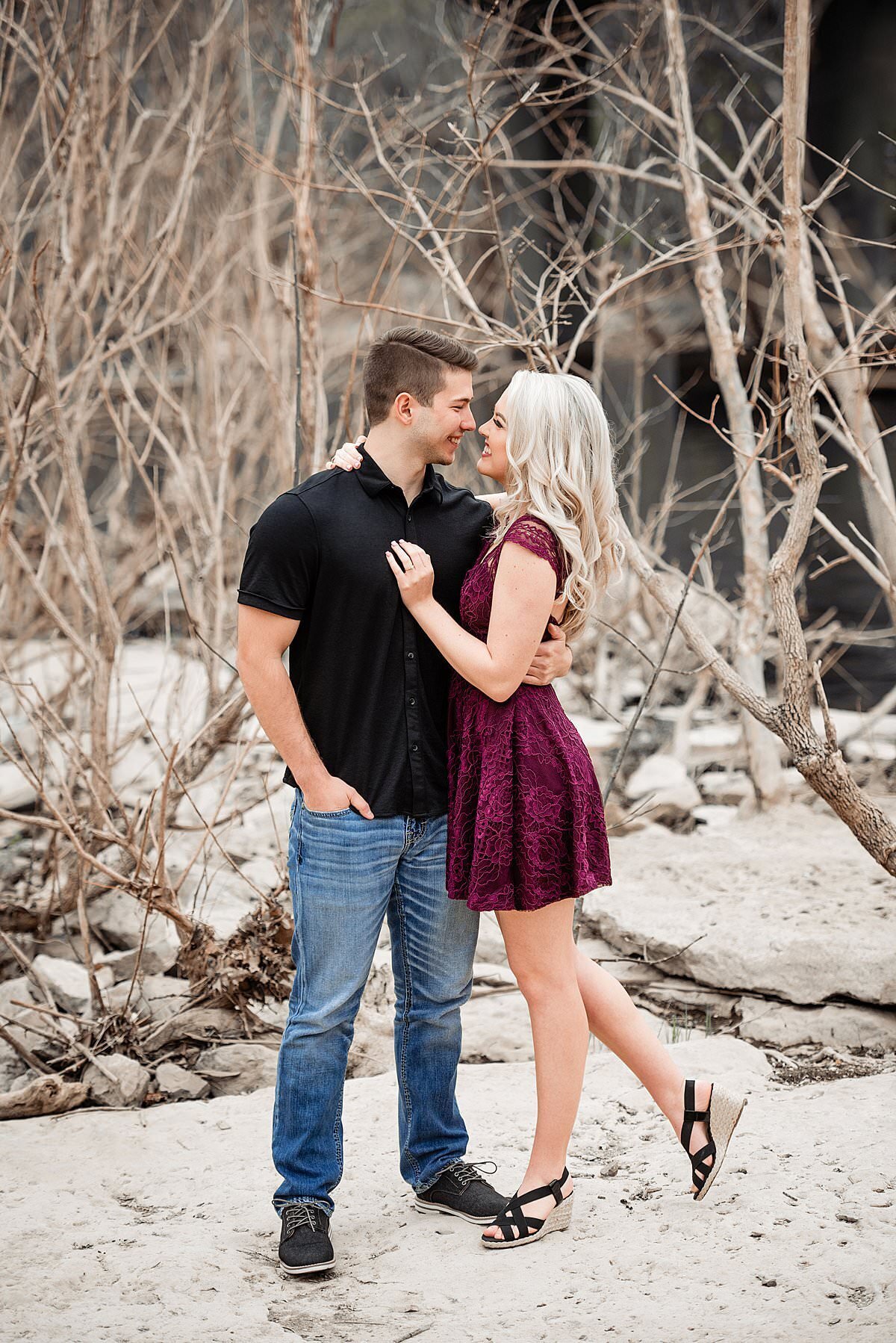 Winter engagement photos taken on Murfreesboro greenway near a river with girl wearing a maroon lace dress and groom a black polo shirt and jeans, they are snuggled together and embracing