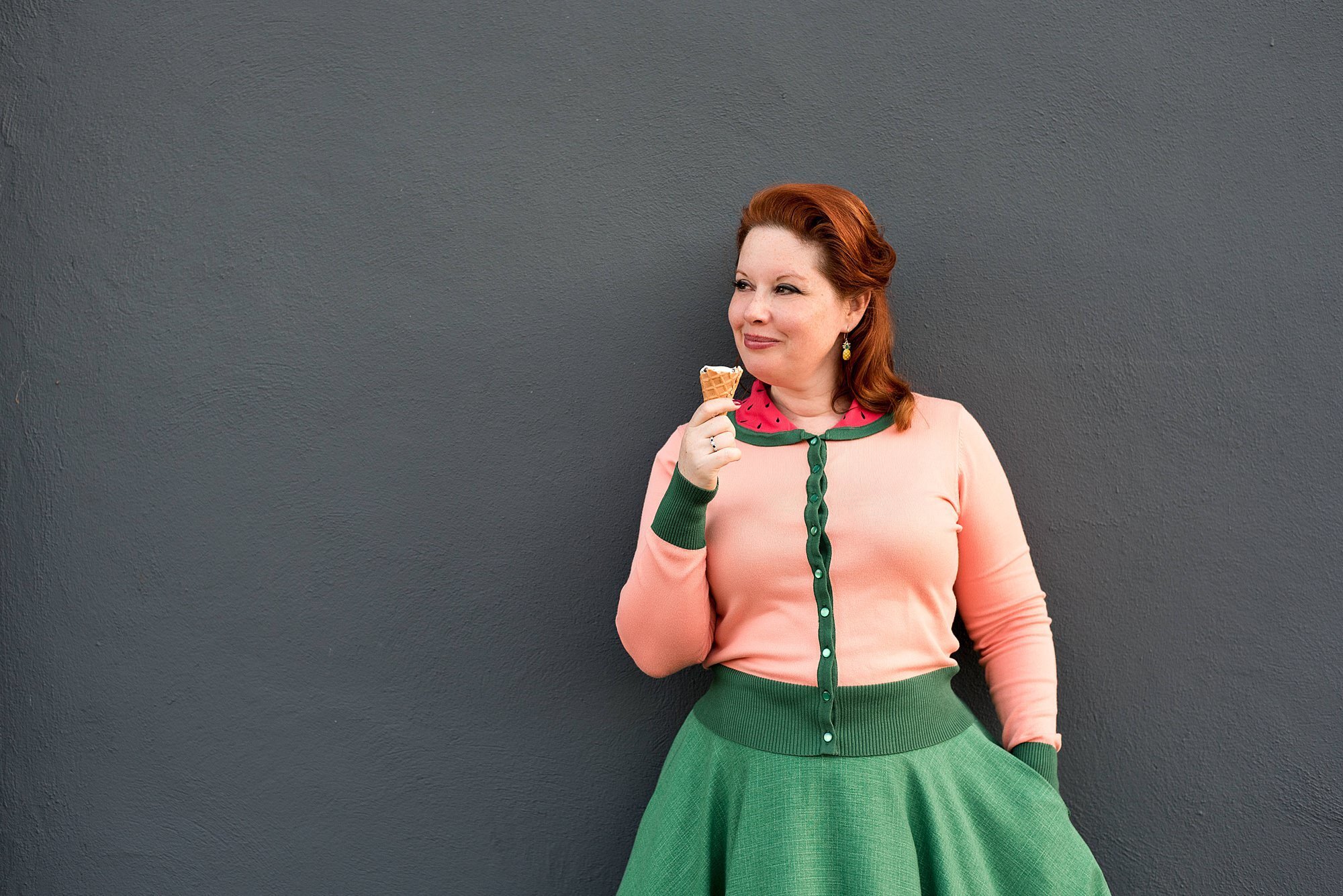 Raina in front of soft blue wall wearing a watermelon inspired skirt and sweater combo eating ice cream from Hattie Jane's on the square in Murfreesboro