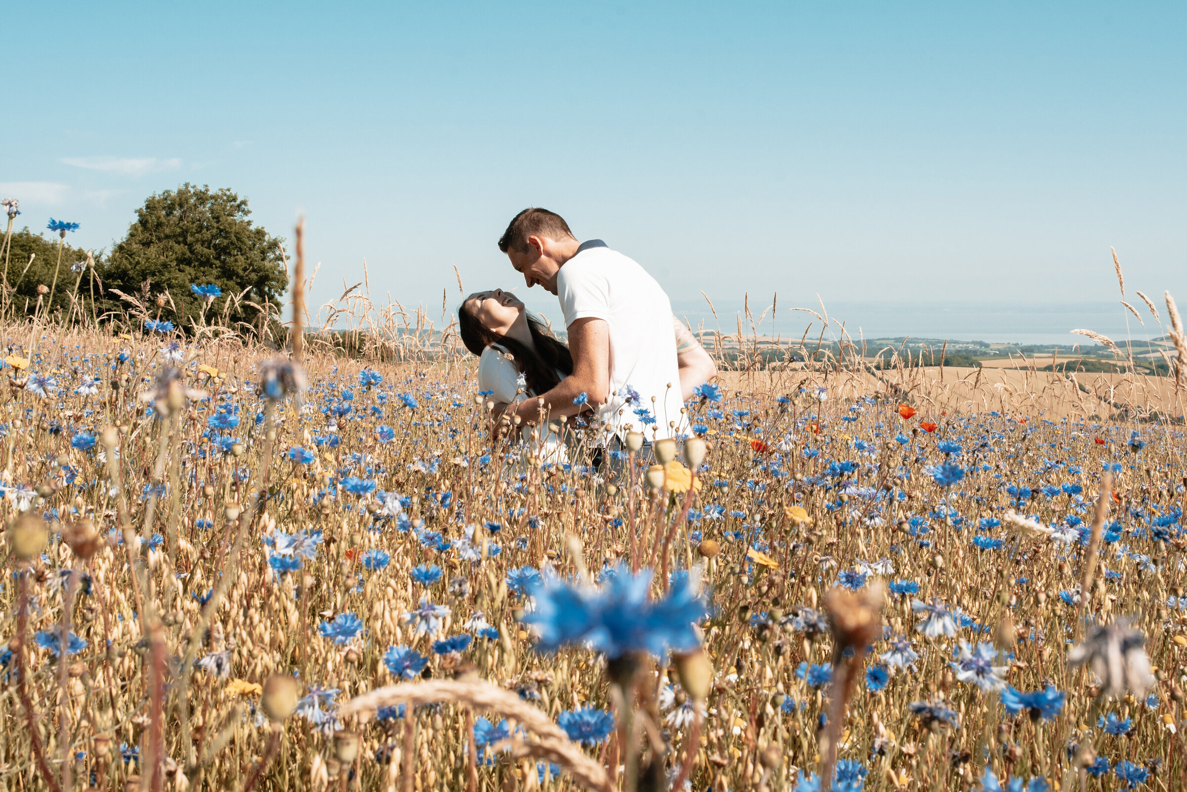 Man tickling woman so much that she is bending backwards and laughing, in a wildflower meadow surrounded by flowers