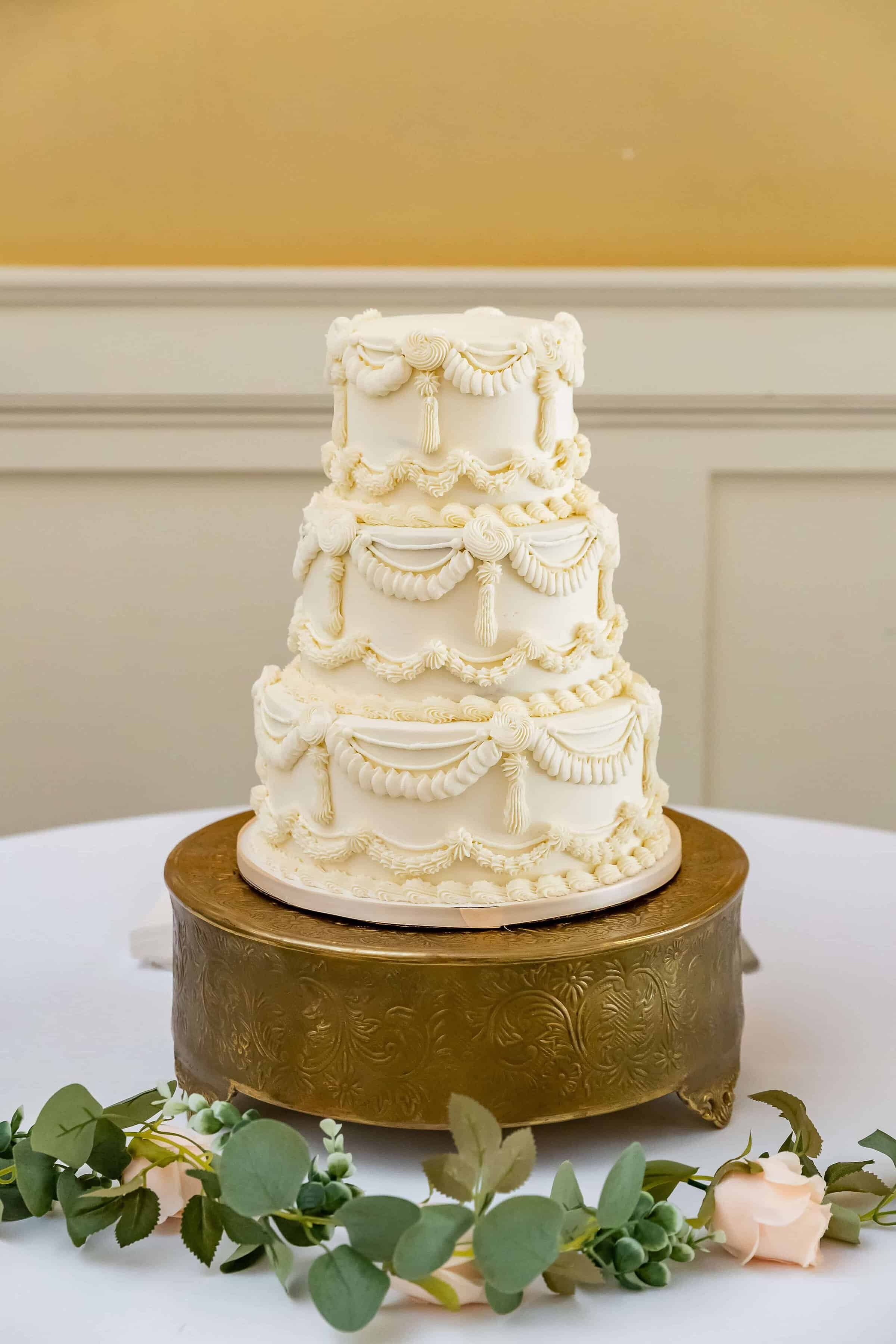 A three-tiered white wedding cake with ornate icing decorations sits on a gold cake stand, placed on a white table with green eucalyptus leaves and a pale pink rose.