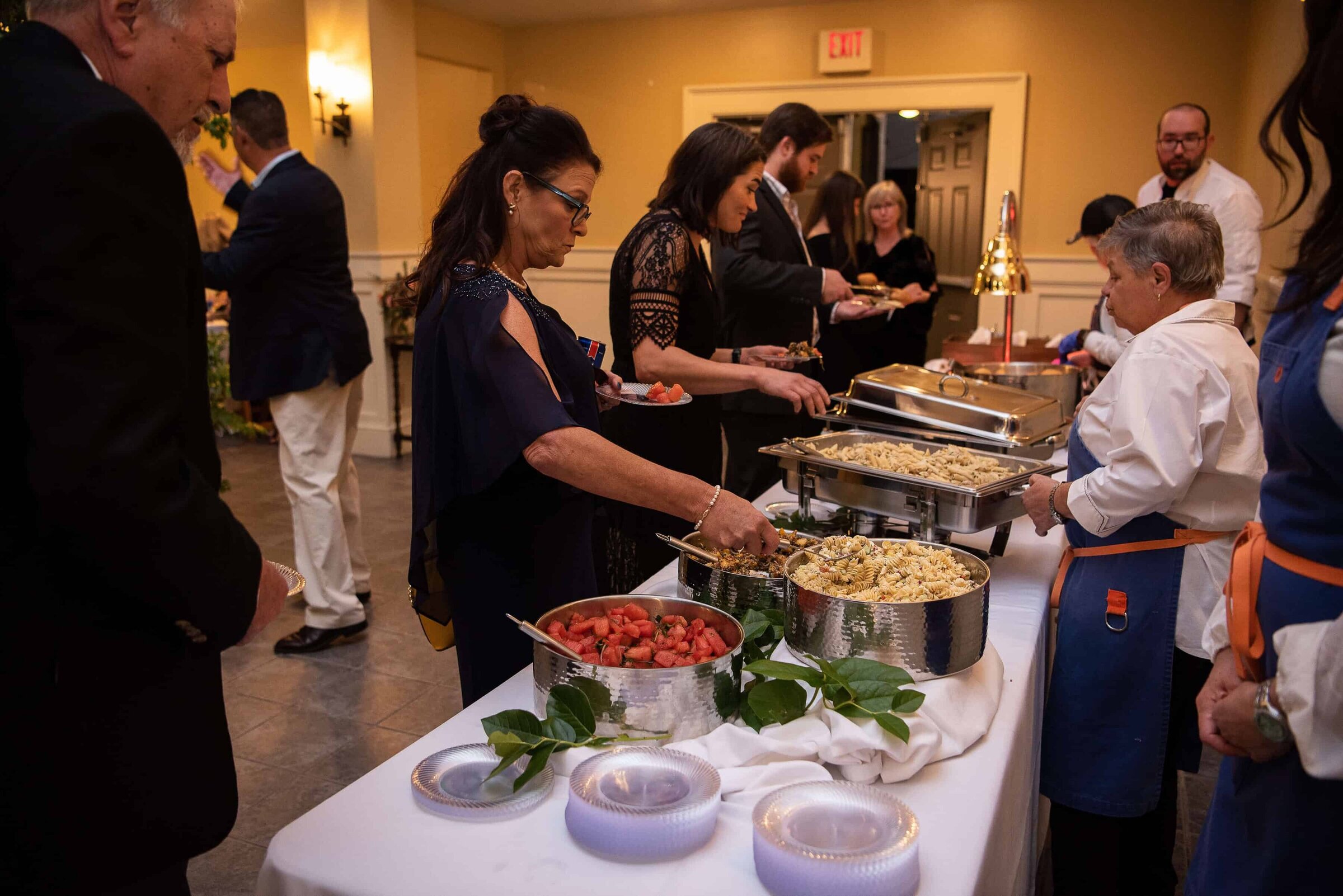 People serve themselves food from a buffet table at a private event, with trays of pasta and a large bowl of fruit salad.