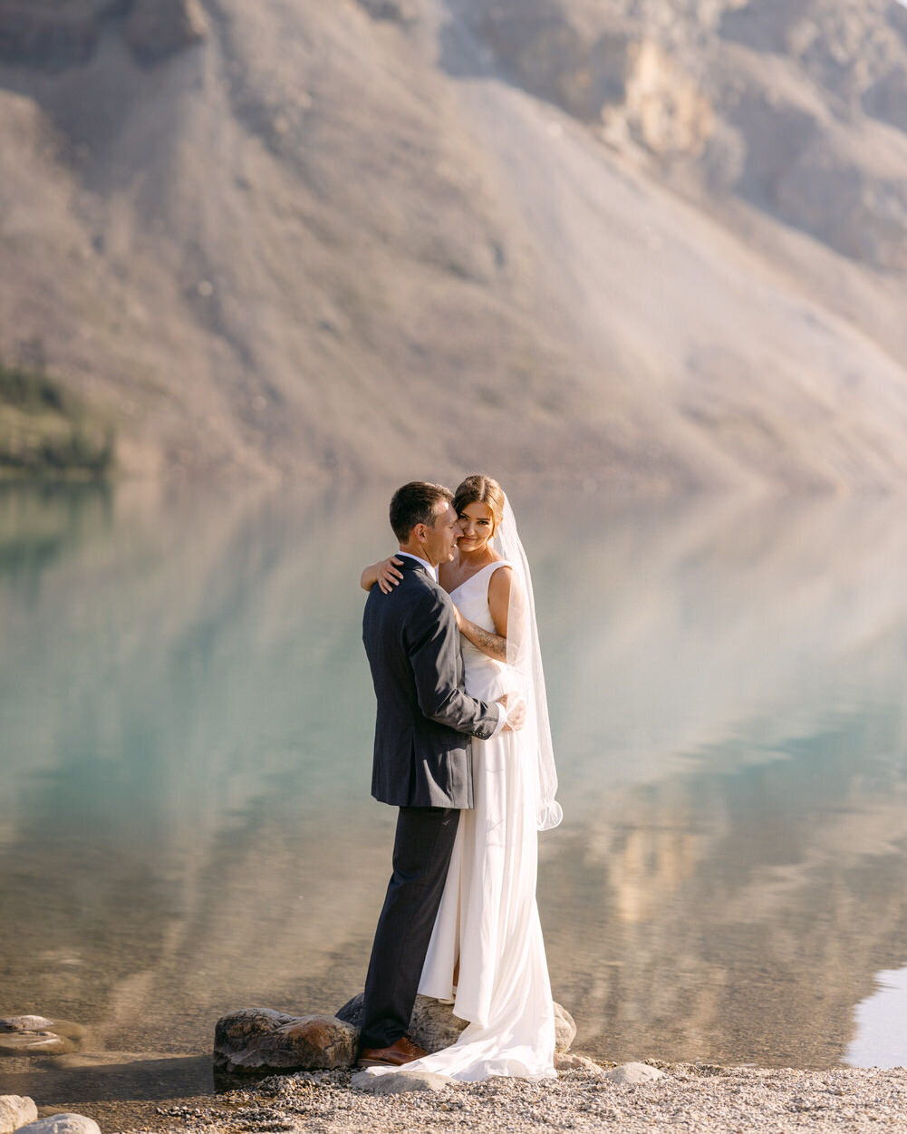 This captivating image captures the bride and groom holding each other by Bow Lake at sunset. The rare calm of the lake's still waters reflects the breathtaking scenery, creating a peaceful and romantic moment as the couple enjoys the tranquil beauty of their wedding day.