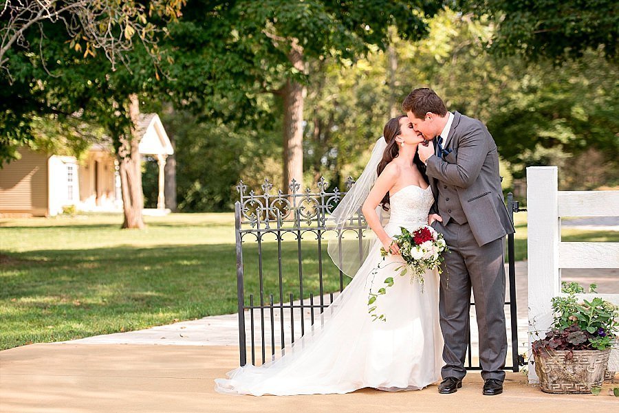 Summer day, groom wearing a grey suit tilting his brides chin up to give her a kiss while standing next to a white fence and ironwork gate
