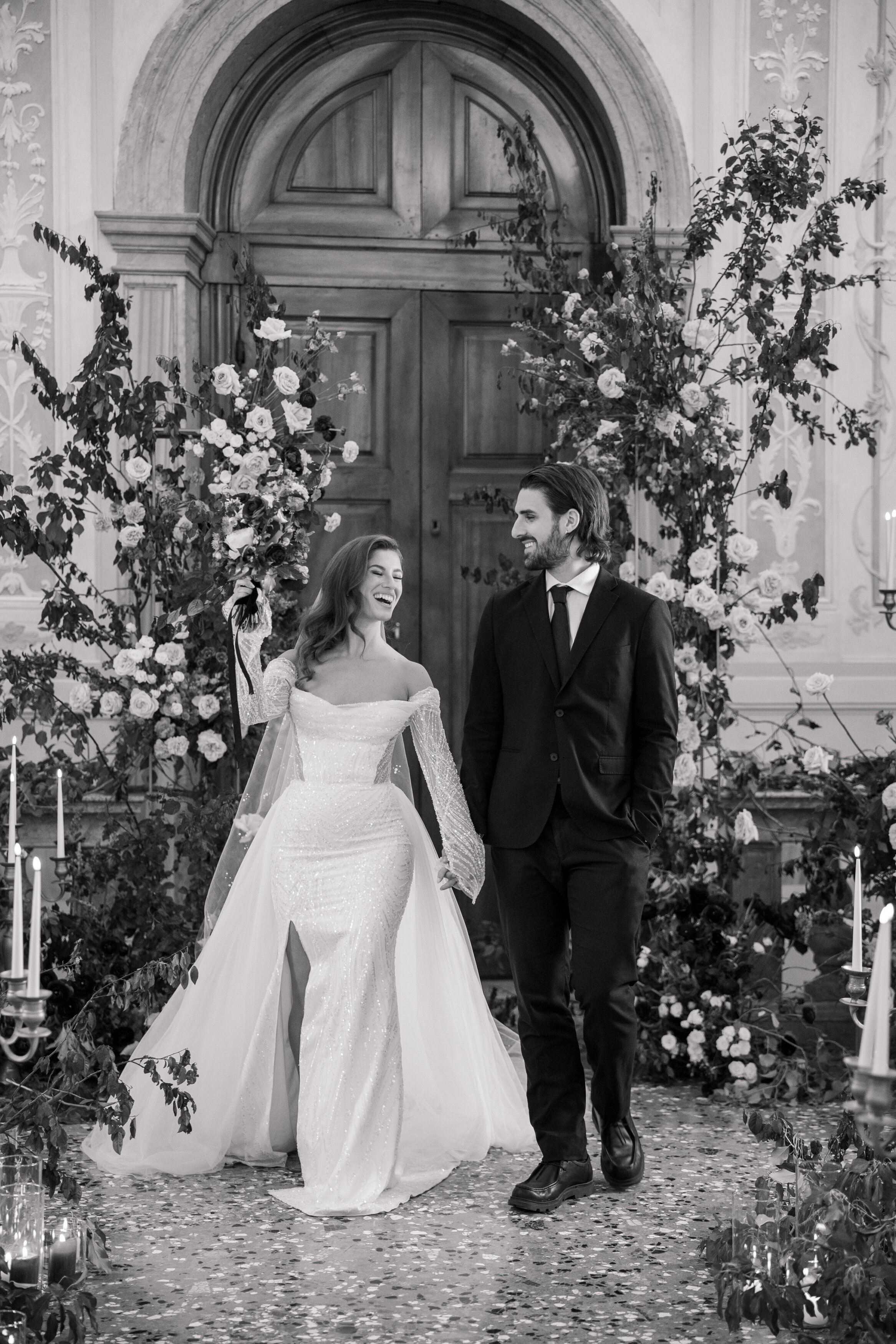 Wedding and Elopement Photographer, black and white image of couple smiling in front of large wooden door and flower arrangements