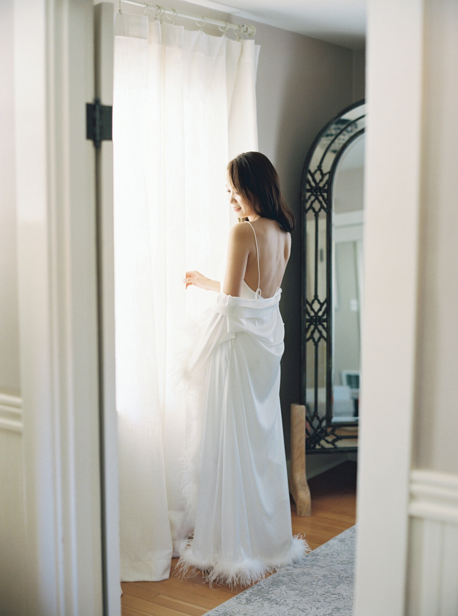 bride in white silk robe looking out window