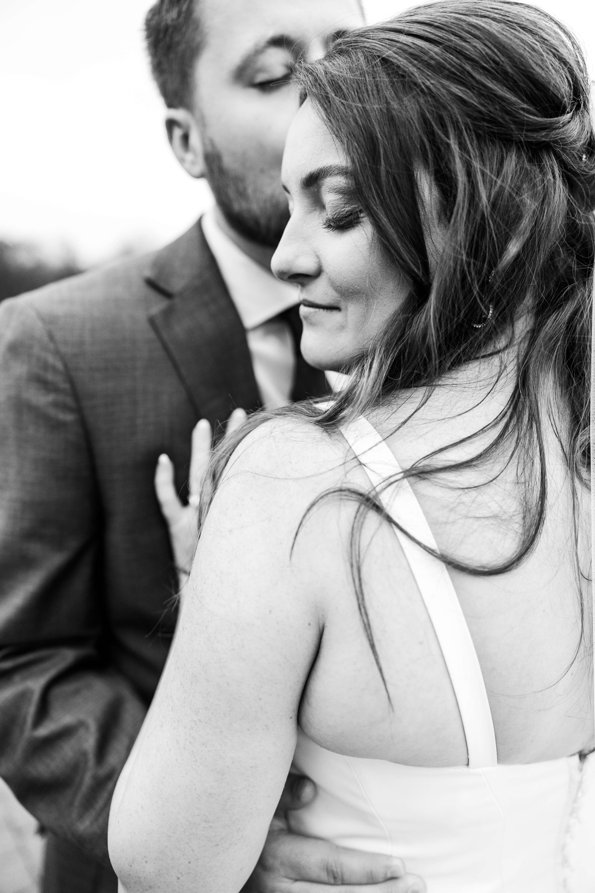 A bride stands with her back to the camera looking over her left shoulder with her groom giving her a kiss on the cheek. It is a black and white photo taken at cooper creek event center.