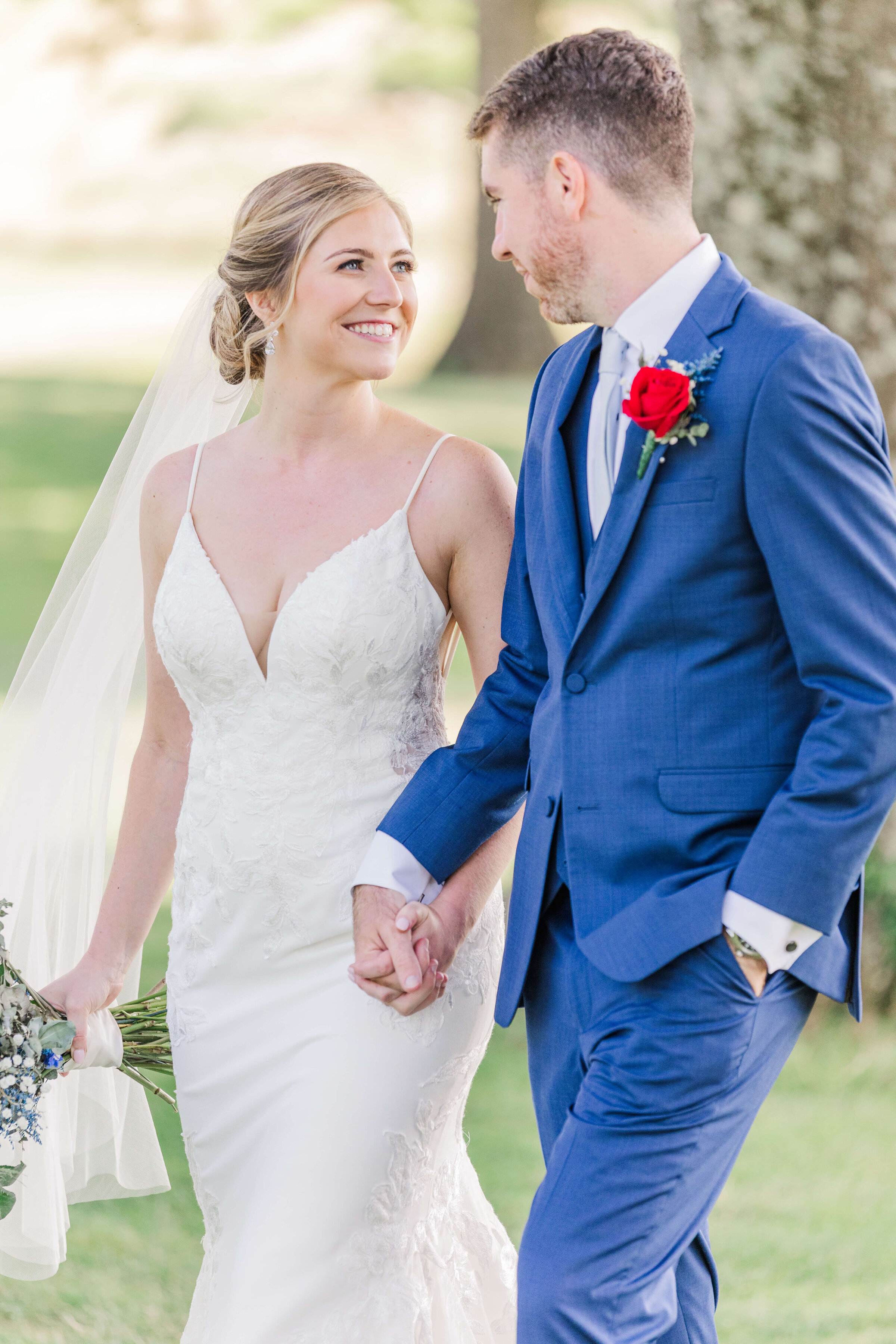 A bride and groom walk and hold hands looking at each other. Around them the grass and trees are really green. He's wearing a blue tux and she's holding a red, white, and blue bouquet