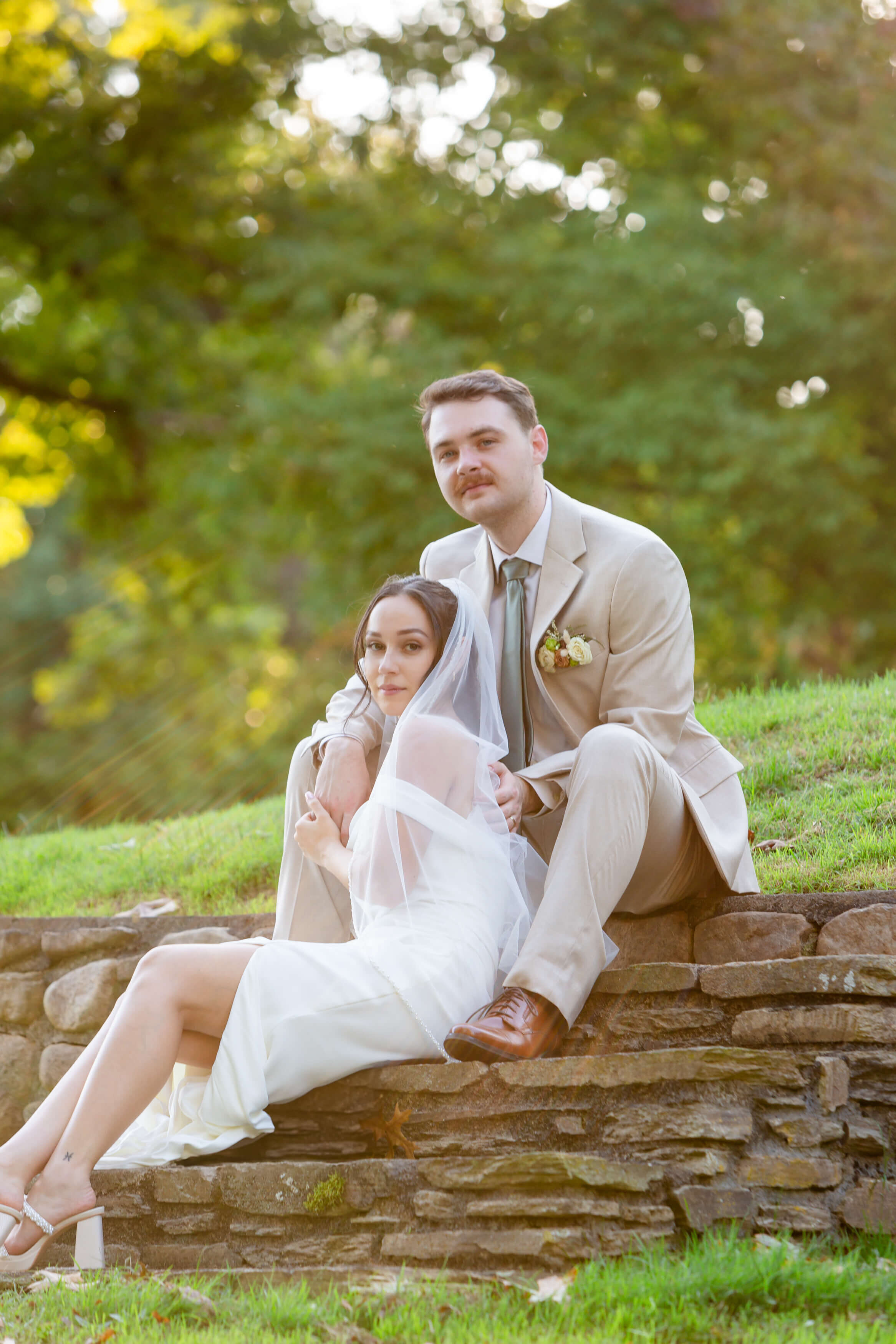 Romantic newlyweds seated on rustic stone steps, the bride draped in a soft veil and the groom in a tailored tan suit, capturing an intimate, upscale wedding moment in Asheville, NC