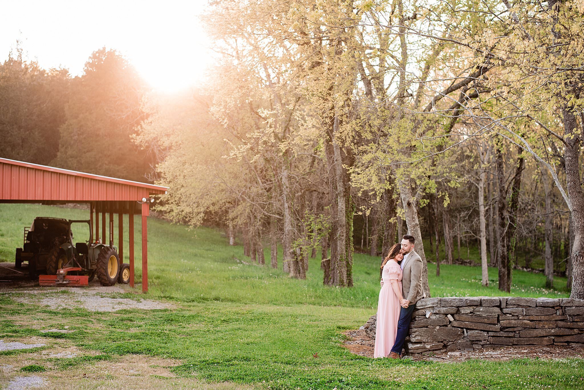 Engagement photo of couple leaning against rock wall with open field and trees in the background and sun setting behind the treeline
