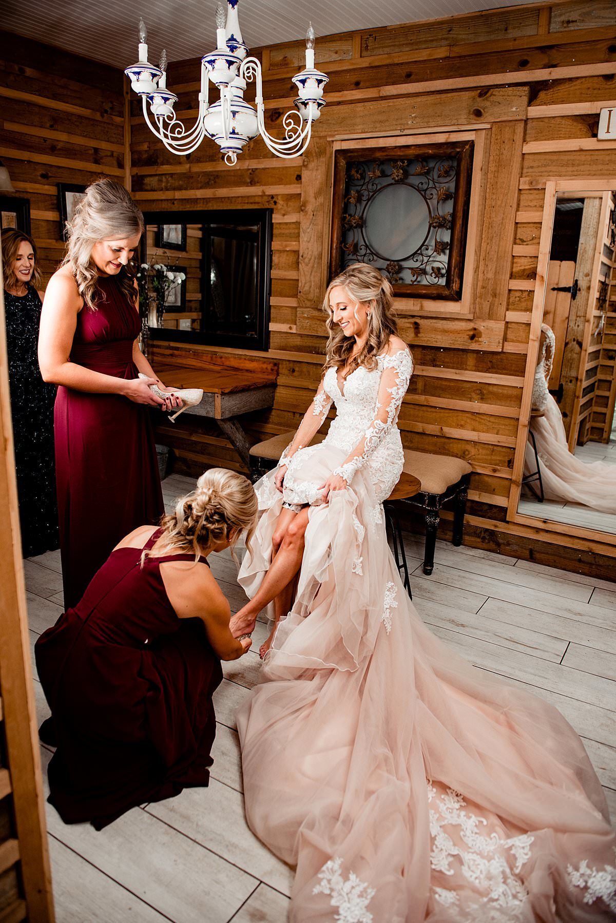 Bridesmaids helping bride put her garter on, wood getting ready room with white chandelier