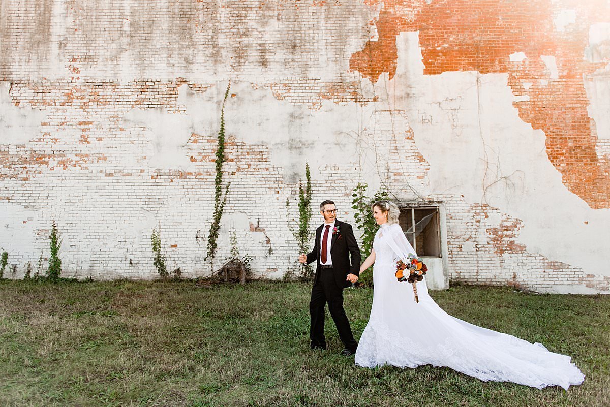 Bride wearing her mothers wedding dress and walking with her husband in front of a white and brick distressed wall